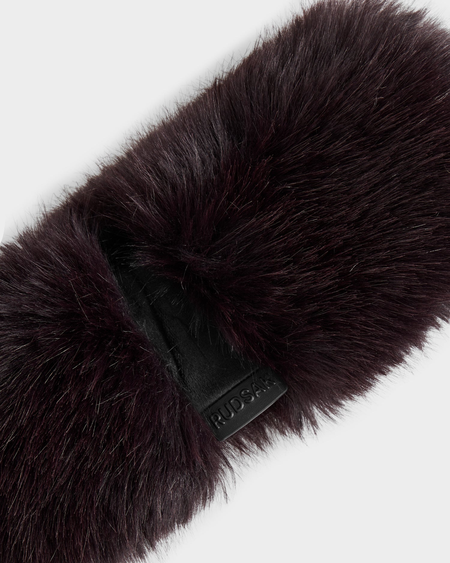 A close-up of the ARLETTE - 8425544 Bordeaux women’s faux fur headband by Rudsak, featuring a black leather tag with embossed branding, set against a light background—an ideal cozy-chic winter accessory.