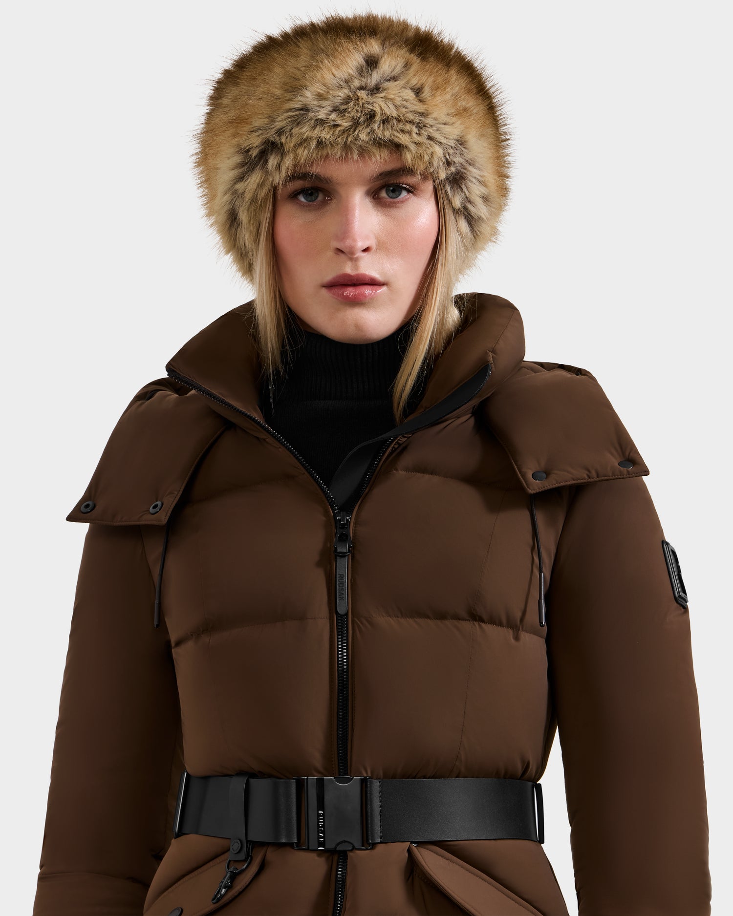 A woman styles the Rudsak ARLETTE - 8425544 Mix Mocha puffer coat, featuring a black belt and fur-lined hood. She pairs it with straight blonde hair, a neutral expression, and a women's faux fur headband for a cozy-chic look.