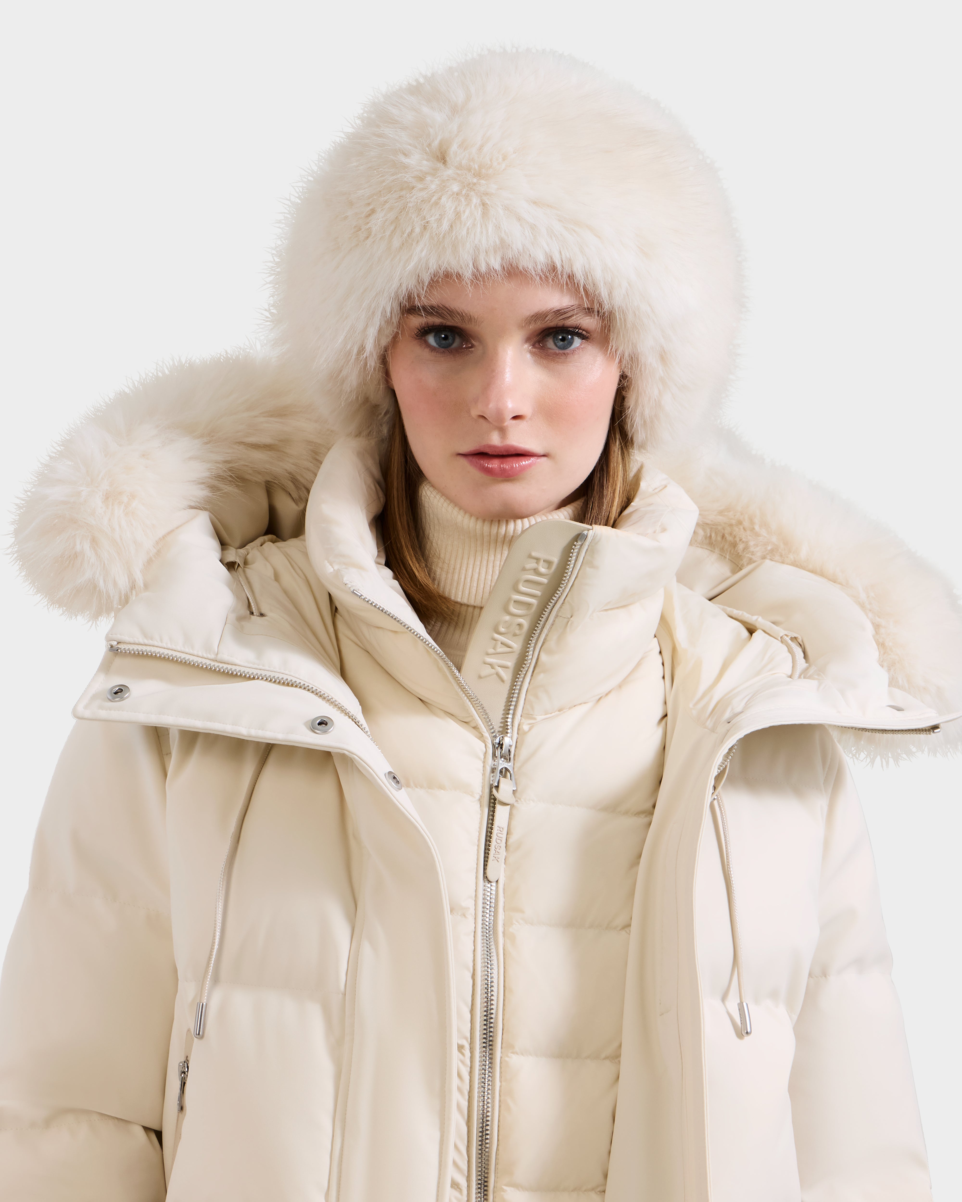 A woman wearing the Rudsak ARLETTE - 8425544 Vanilla puffer jacket with a large fur-trimmed hood and a chic faux fur headband, looking directly at the camera against a plain light background.