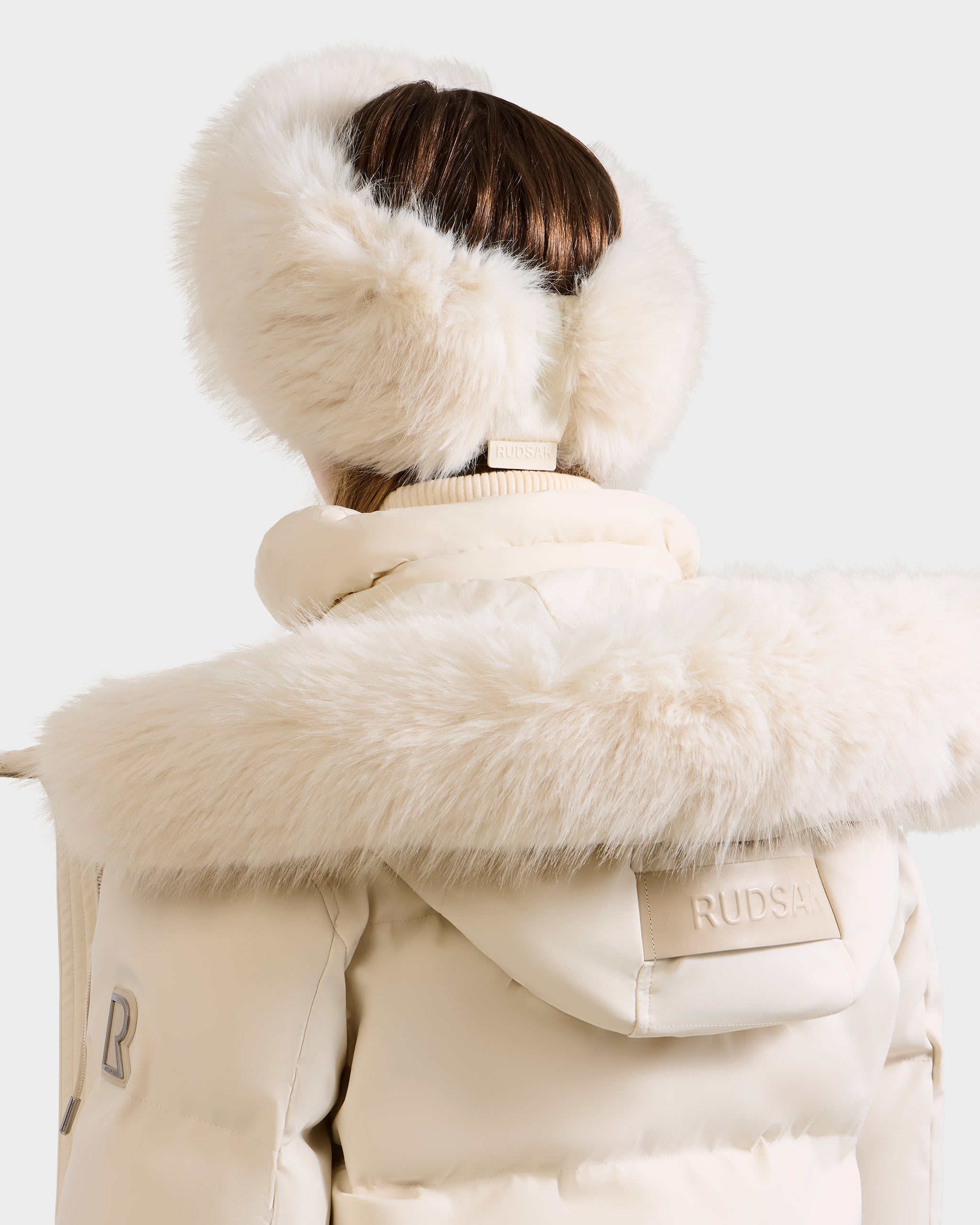 A person seen from behind wears the Rudsak ARLETTE - 8425544 Vanilla coat with a fur-trimmed hood and matching earmuffs, featuring a Rudsak patch on the back.