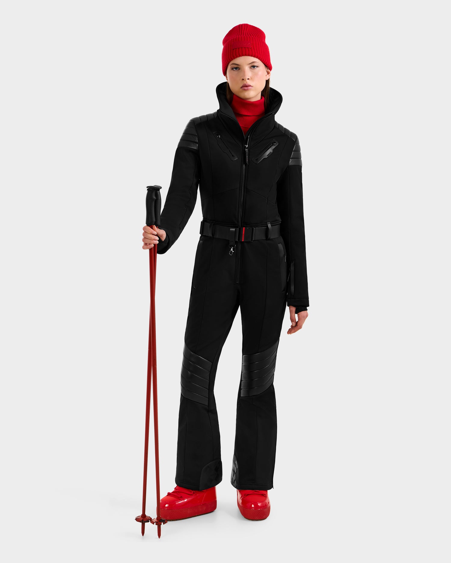Wearing the Rudsak ARLEY - 8124717 Black one-piece ski suit, a person stands with red ski poles, red beanie, and red ski boots against a plain light background.
