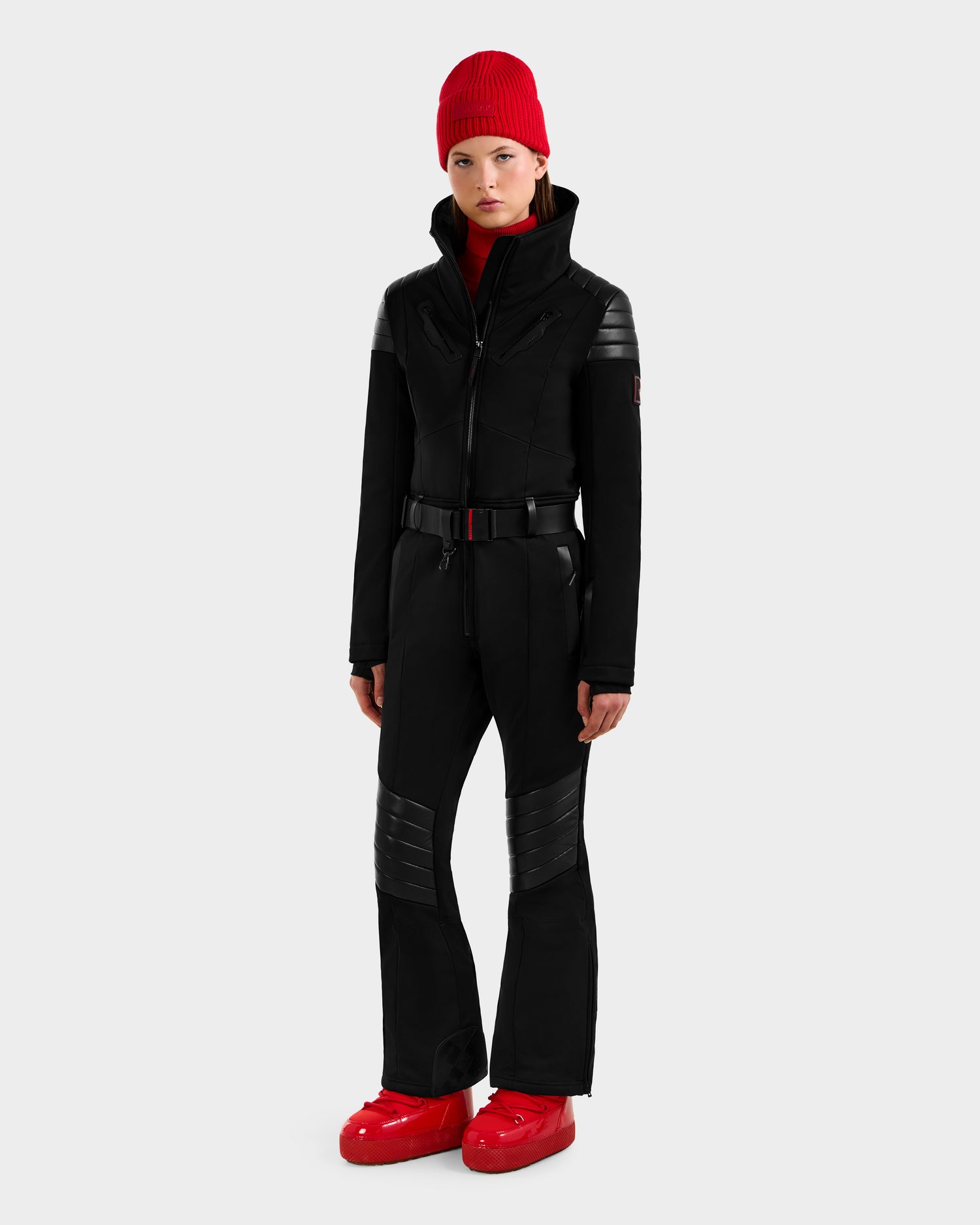 A person in the Rudsak ARLEY - 8124717 Black fitted one-piece ski suit with quilted details, paired with a red knit beanie and red snow boots, stands against a plain light gray background.