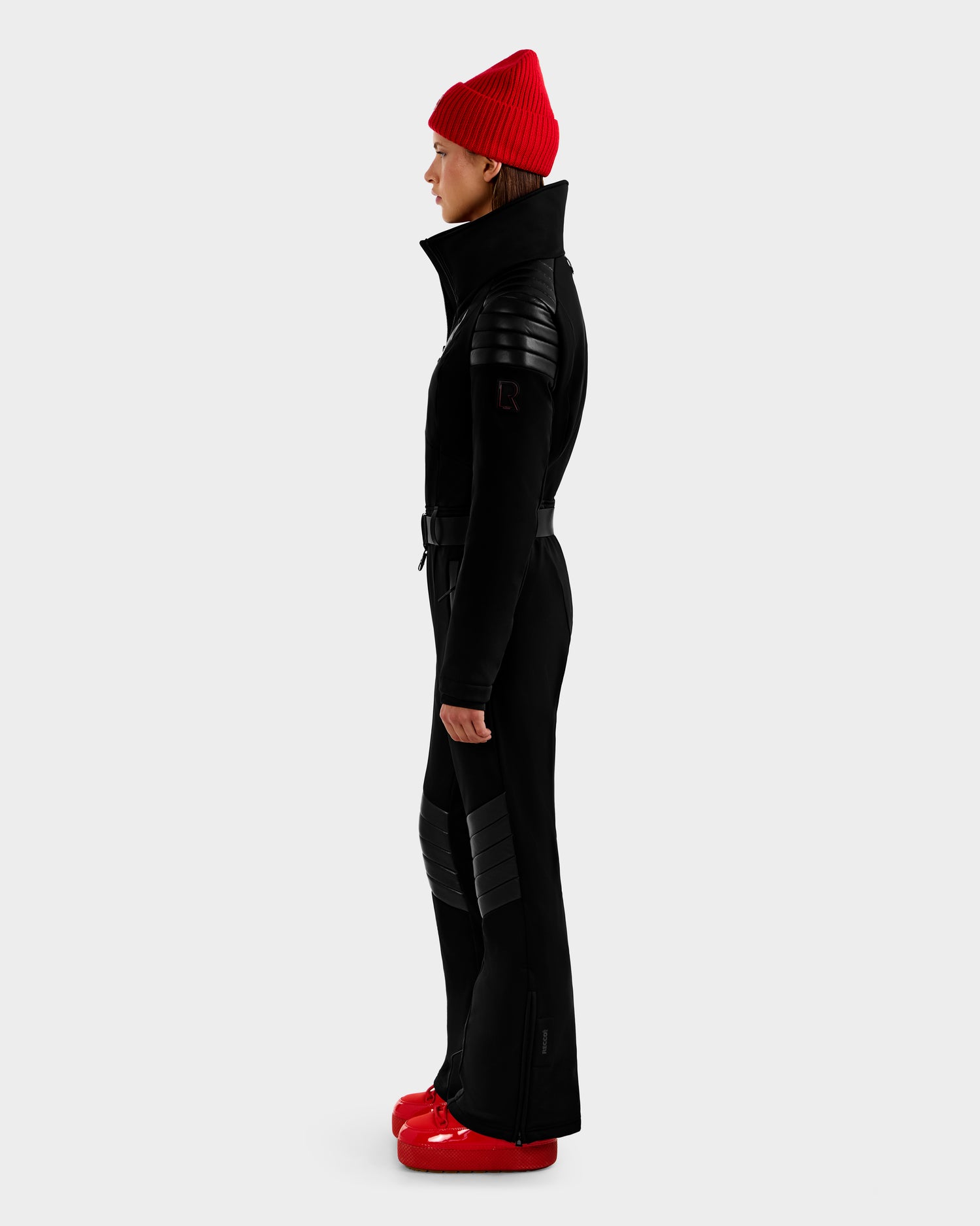 A person in profile wears the Rudsak ARLEY - 8124717 Black ski suit with quilted details and a high collar, paired with a red knit beanie and bright red boots, standing against a plain light gray background.
