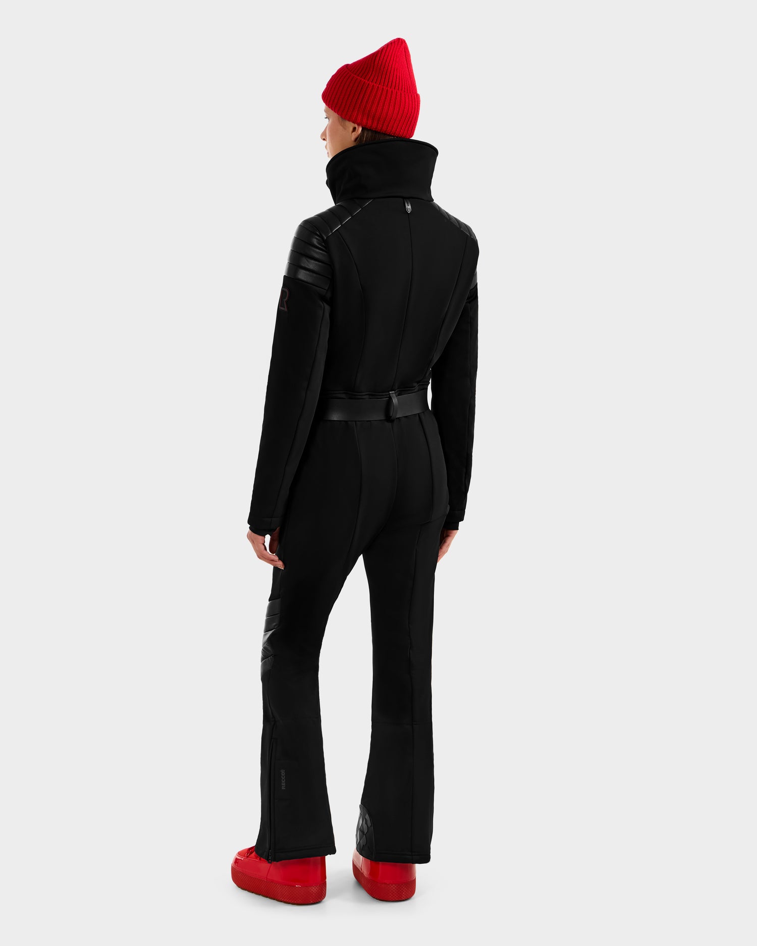 A person stands facing away in the Rudsak ARLEY - 8124717 Black women’s ski suit with a high collar, black belt, and flared legs, paired with a red beanie and red shoes. The background is plain light gray.