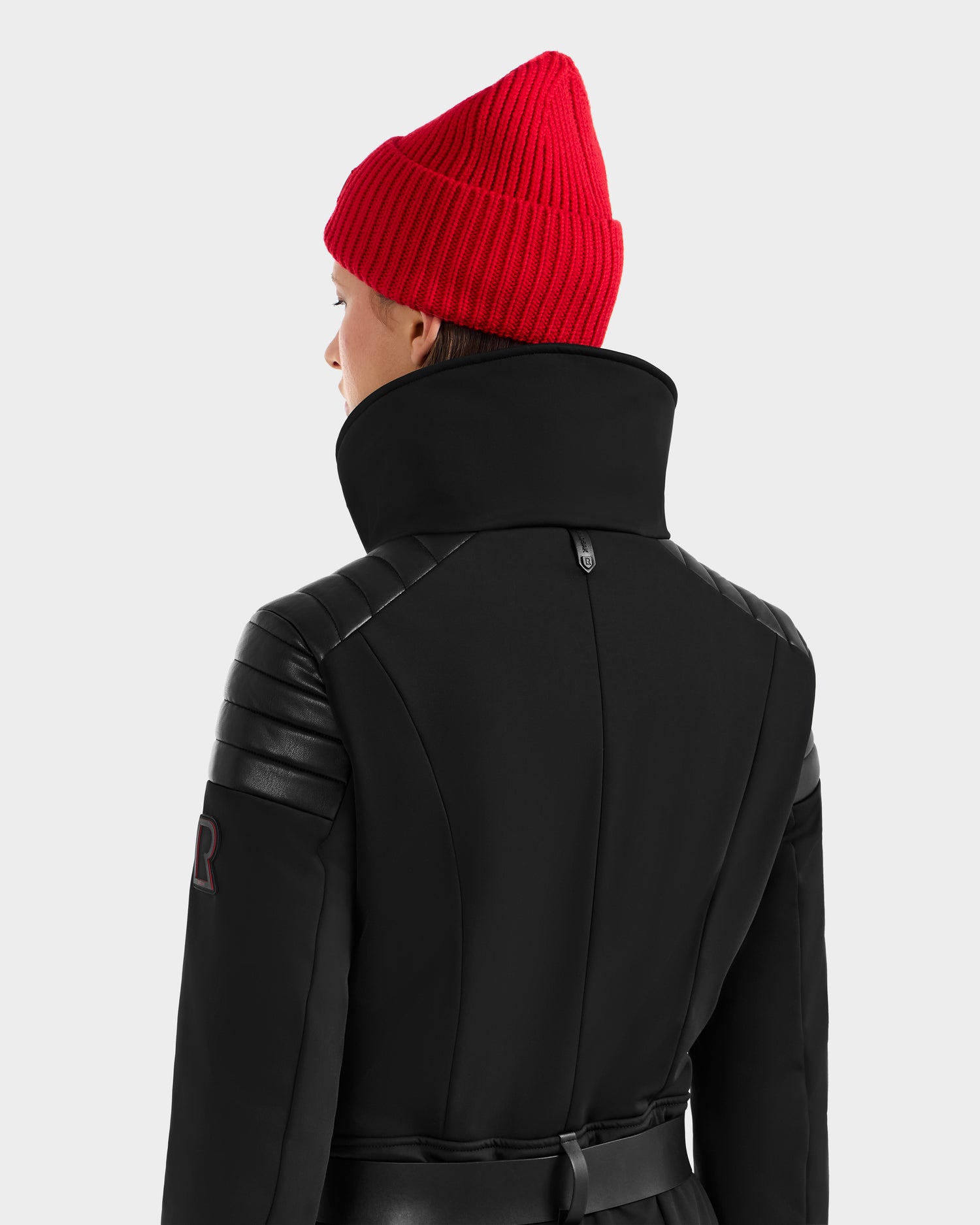 Someone wears the Rudsak ARLEY - 8124717 Black high-collared coat with leather accents and a bright red knit beanie, photographed from behind against a plain light background.