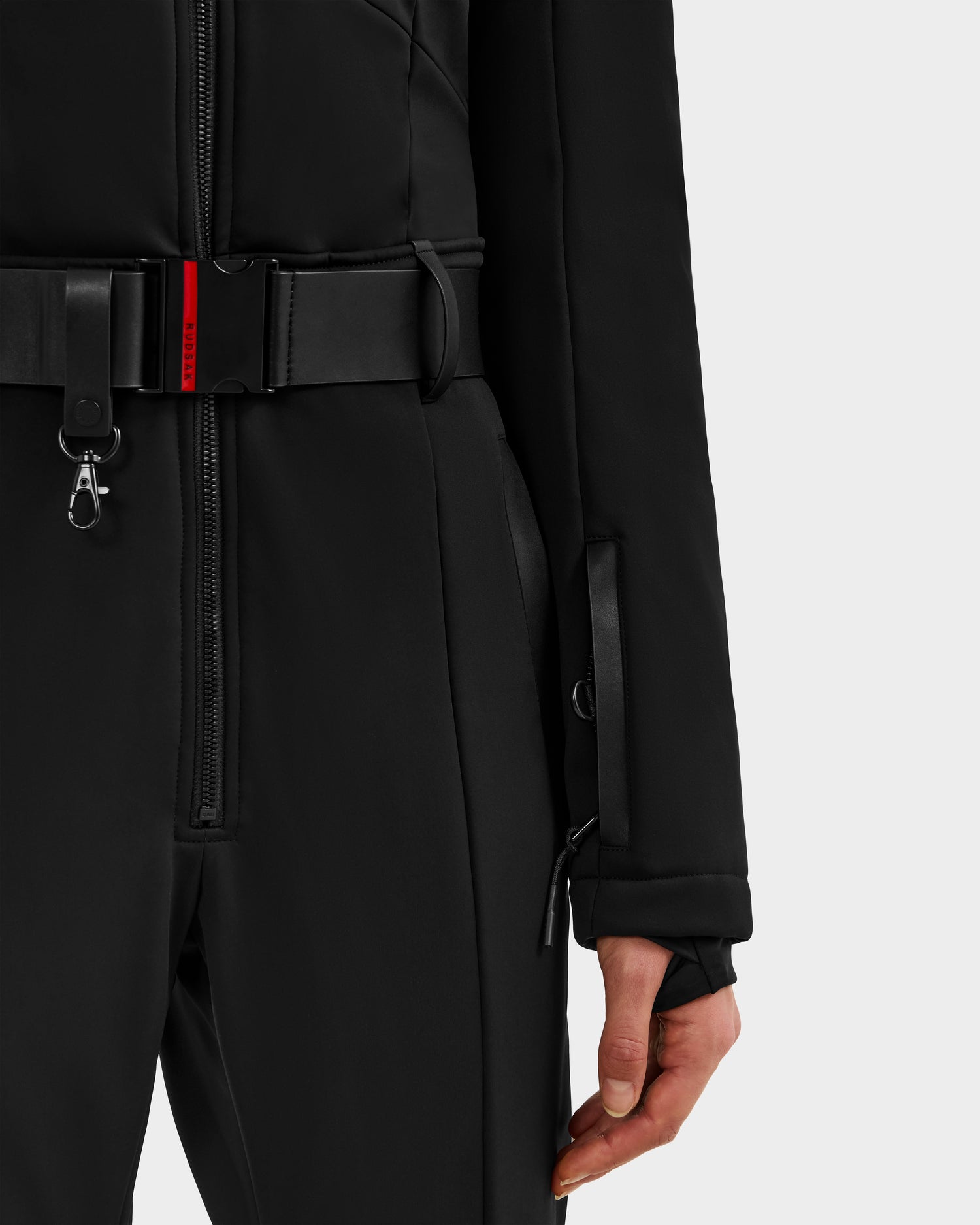 A close-up of a person wearing the Rudsak ARLEY - 8124717 Black women’s ski suit, featuring sleek lines, black zippers, a belted waist, thumb-hole sleeves, and RECCO rescue technology for added safety.
