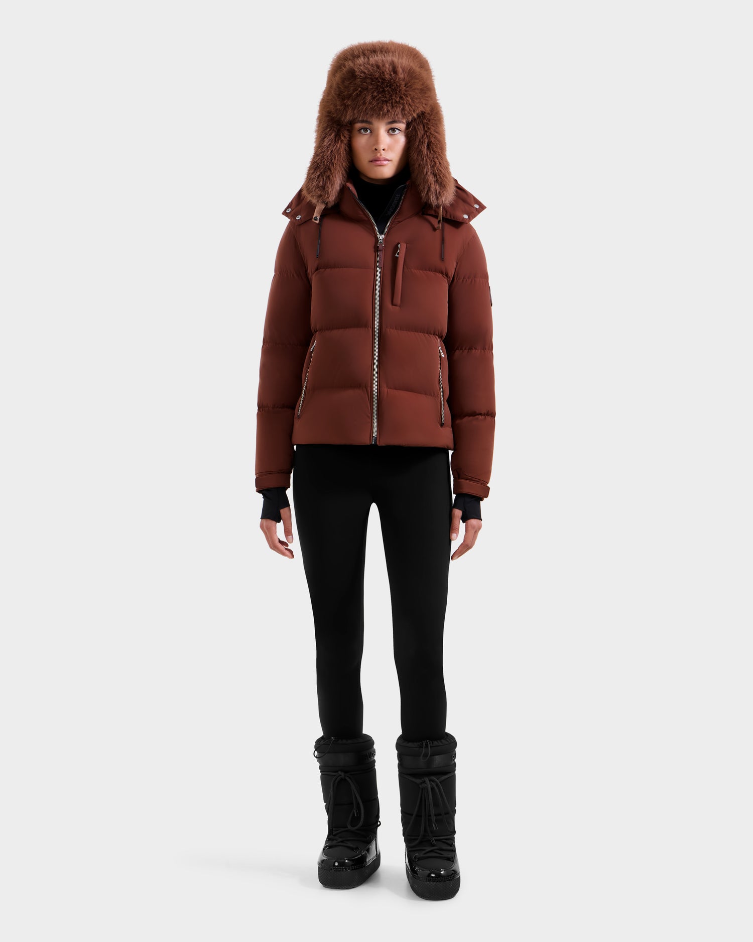A person wears the Rudsak ASHLYN M - 8124564 Rust women's down puffer with a water-resistant shell and fur-lined hood, paired with black leggings and winter boots, standing against a plain white background.