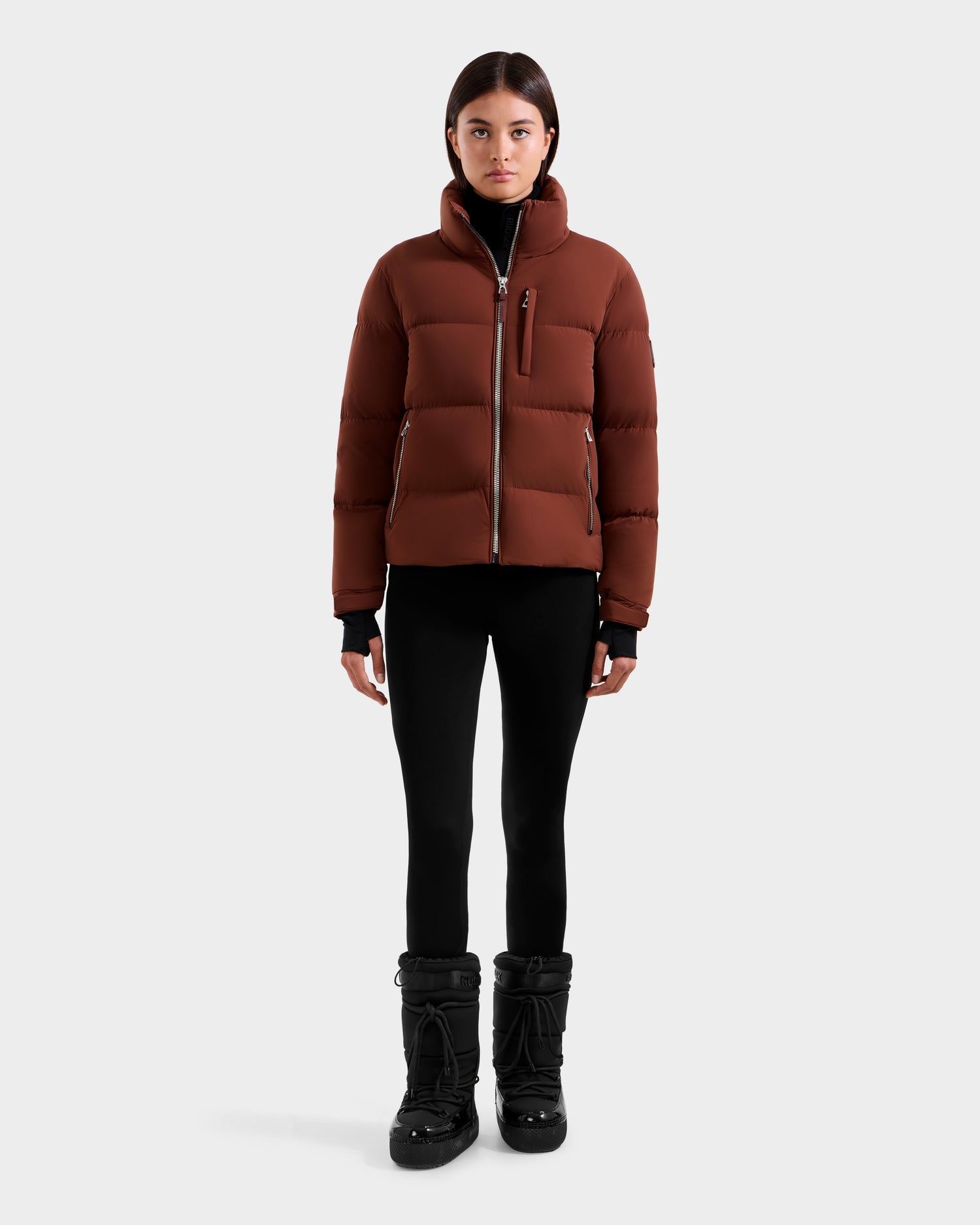 A person stands facing forward, wearing a Rudsak ASHLYN M - 8124564 Rust puffer jacket, black leggings, and black winter boots against a plain light gray background.