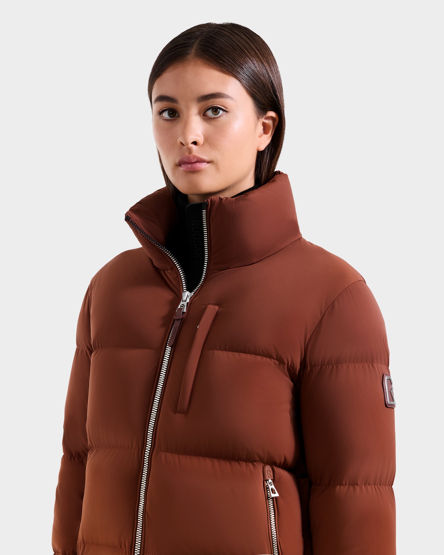 A woman with straight brown hair wears the Rudsak ASHLYN M - 8124564 Rust women’s down puffer jacket with a water-resistant shell and zippered pockets, standing against a plain light gray background.