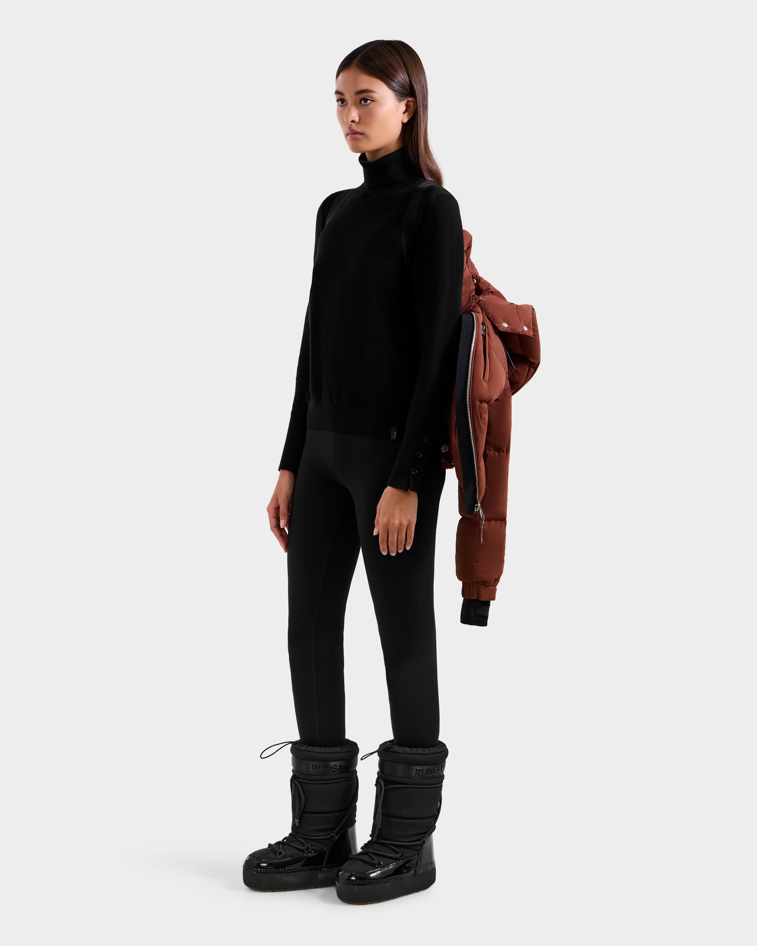 A woman in a black turtleneck, leggings, and winter boots drapes the Rudsak ASHLYN M - 8124564 Rust women's down puffer with water-resistant shell over her left shoulder. Her straight brown hair stands out against a plain light gray background.