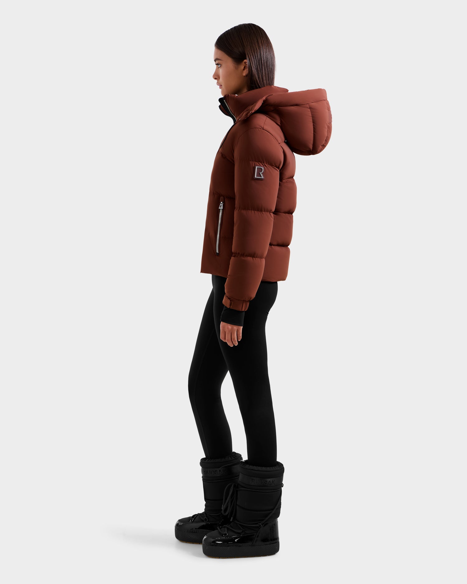 A woman in profile wears the Rudsak ASHLYN M - 8124564 Rust women's down puffer jacket with a hood, paired with black leggings and winter boots, against a light gray background.