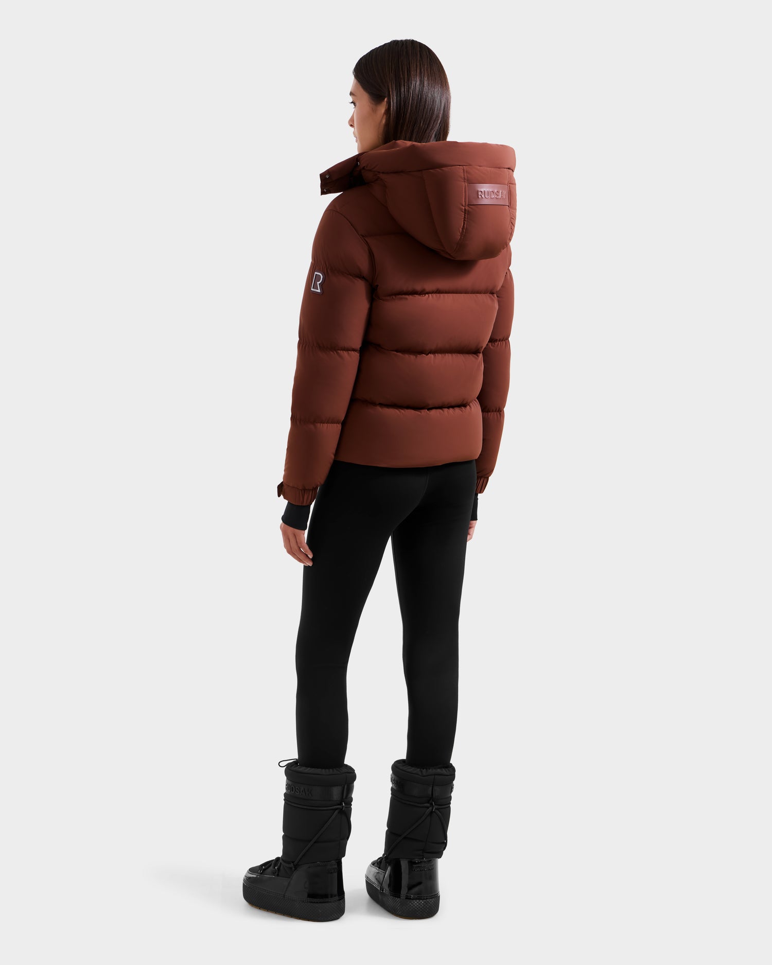 A woman faces away wearing the Rudsak ASHLYN M - 8124564 Rust water-resistant puffer jacket with a hood, black leggings, and winter boots, set against a plain light gray background.