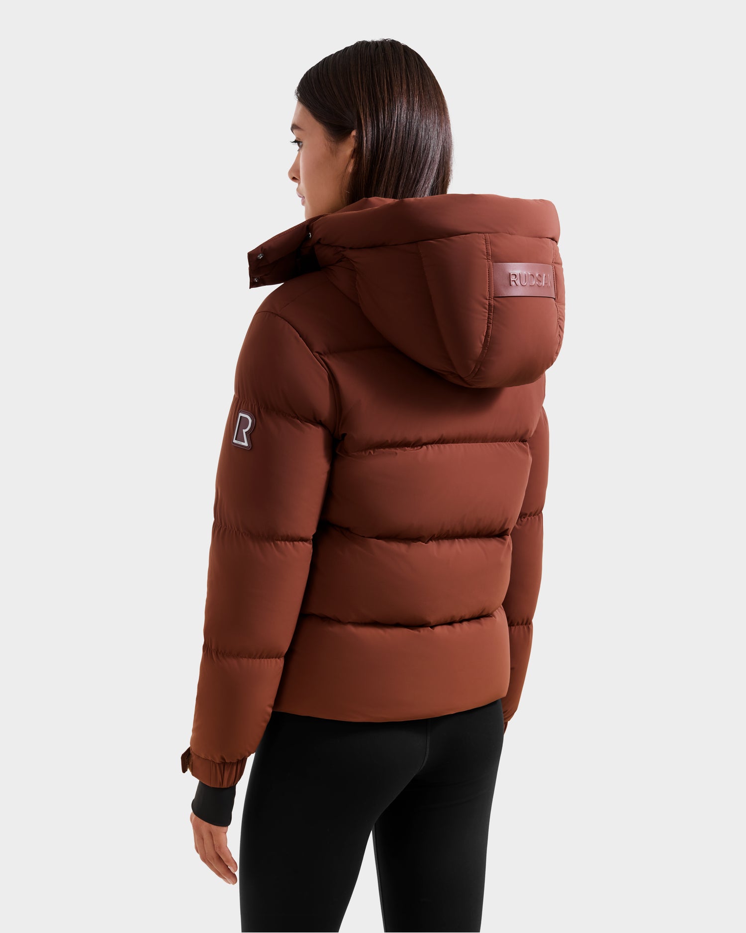 A woman in the Rudsak ASHLYN M - 8124564 Rust down puffer jacket with a water-resistant shell, patch on the sleeve, and logo on the hood stands facing away from the camera, paired with black leggings.