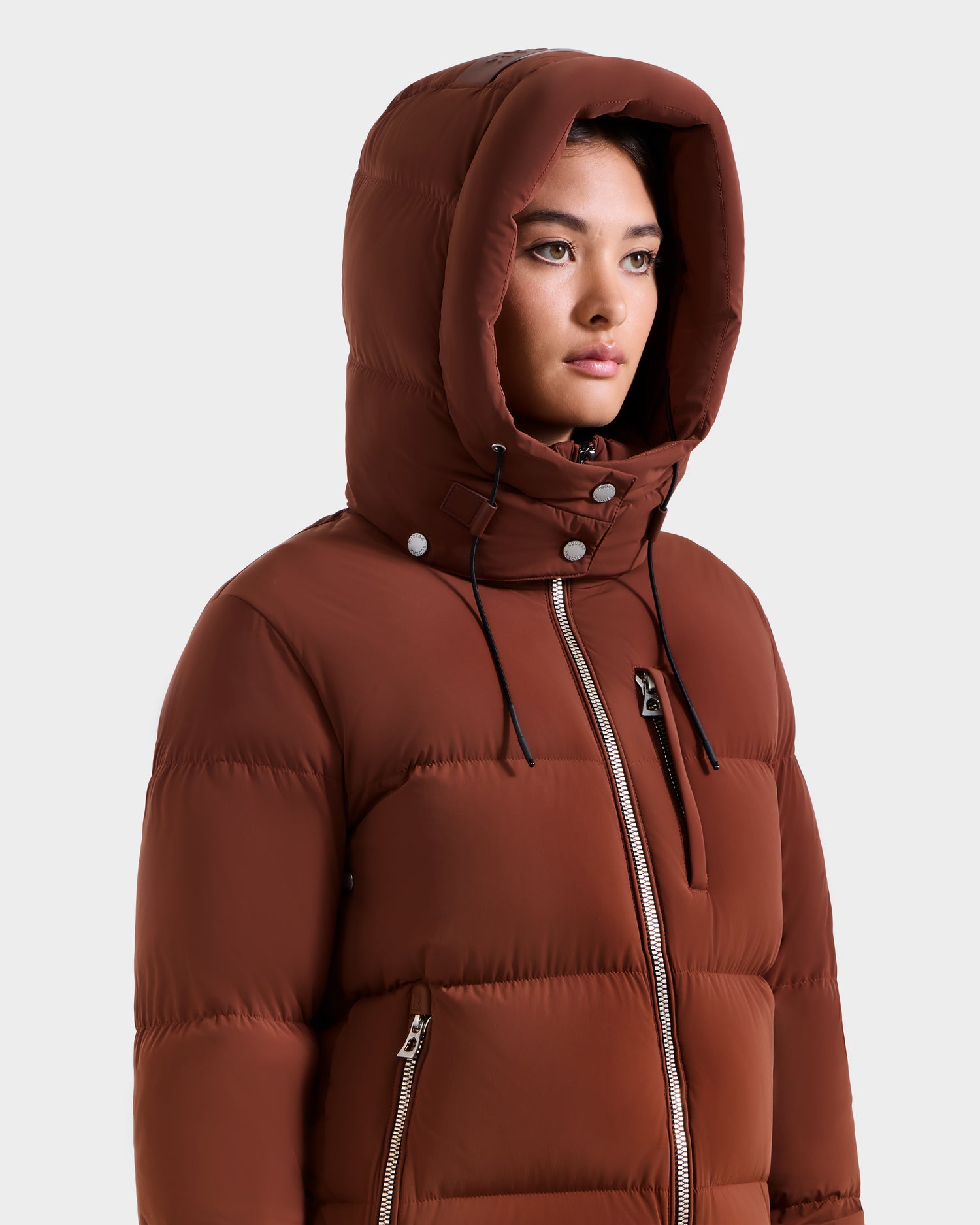 A woman wears the Rudsak ASHLYN M - 8124564 Rust down puffer jacket with hood up, featuring black drawstrings, silver zippers, and a water-resistant shell, against a plain white background.