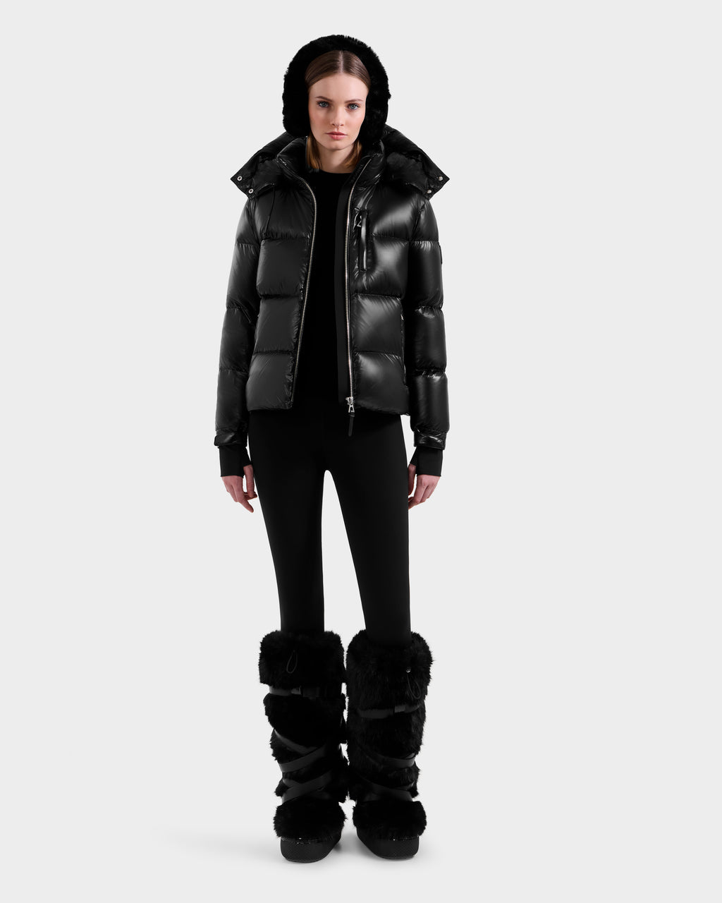 A woman faces forward in a Rudsak ASHLYN S - 8124565 Black water-resistant puffer jacket with a hood, paired with black leggings and tall furry black boots, set against a plain light background.