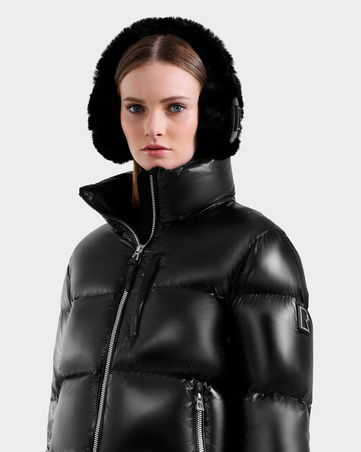 A woman in a shiny black Rudsak ASHLYN S - 8124565 down puffer jacket and large black earmuffs looks ahead with a neutral expression against a plain light background.