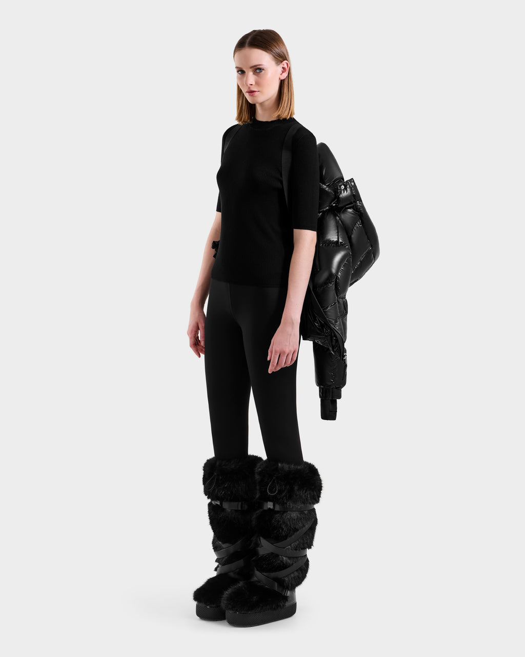 A woman stands against a plain background wearing the Rudsak ASHLYN S - 8124565 Black puffer jacket draped over one shoulder, paired with a black top, black leggings, and black furry boots.