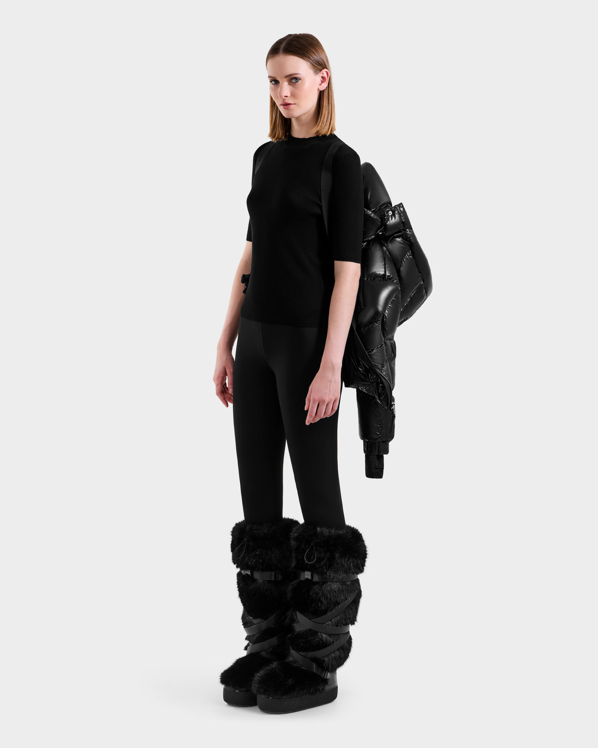 A woman stands against a plain background wearing the Rudsak ASHLYN S - 8124565 Black puffer jacket draped over one shoulder, paired with a black top, black leggings, and black furry boots.