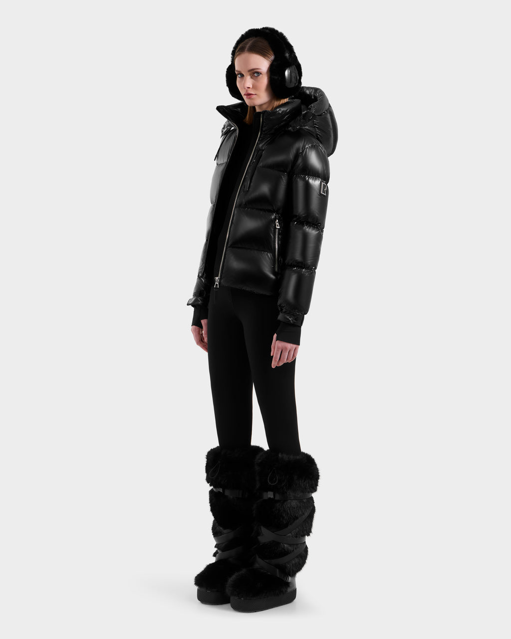 A woman stands against a plain background wearing the Rudsak ASHLYN S - 8124565 Black water-resistant puffer jacket with a hood, paired with black earmuffs, leggings, and tall black fur boots.