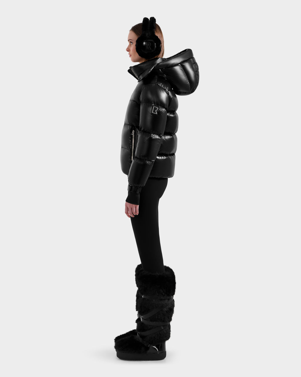 A woman stands in profile wearing the Rudsak ASHLYN S - 8124565 Black down puffer jacket with a hood, black leggings, furry boots, and earmuffs against a plain light gray background.