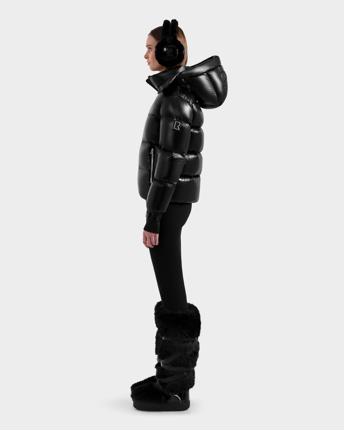 A woman stands in profile wearing the Rudsak ASHLYN S - 8124565 Black down puffer jacket with a hood, black leggings, furry boots, and earmuffs against a plain light gray background.