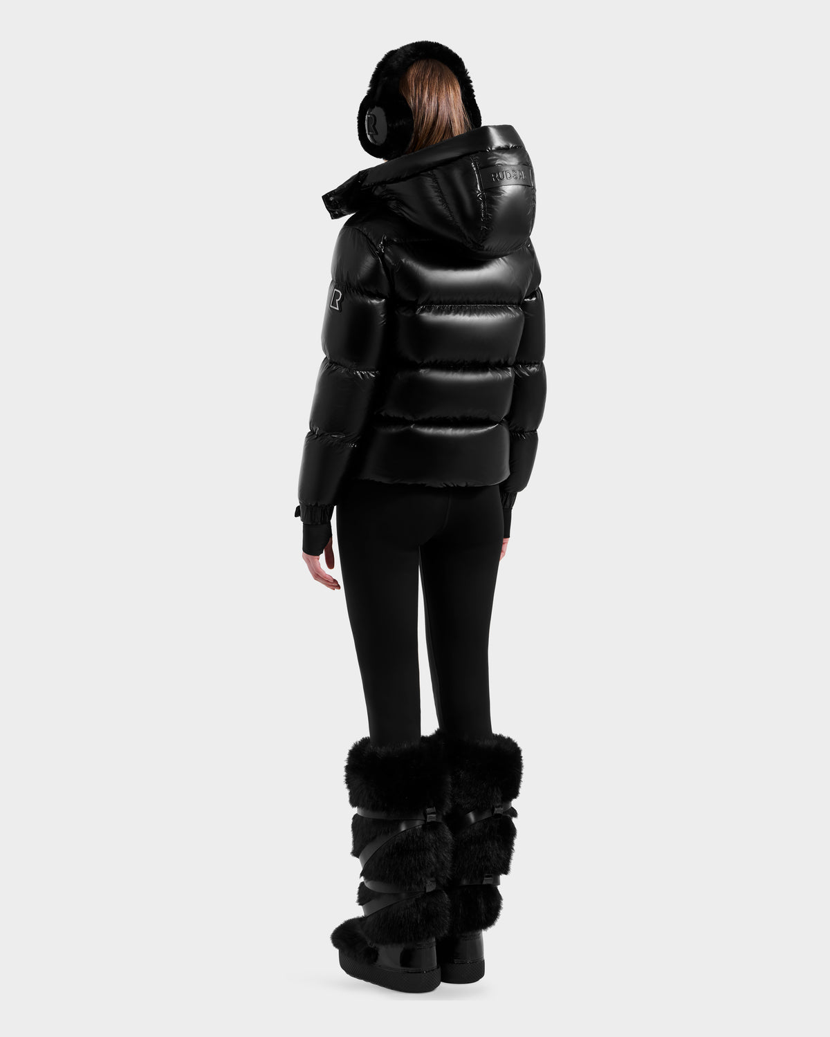 A person faces away wearing the Rudsak ASHLYN S - 8124565 Black water-resistant puffer jacket with a hood, paired with black leggings, furry black boots, and earmuffs against a plain light background.