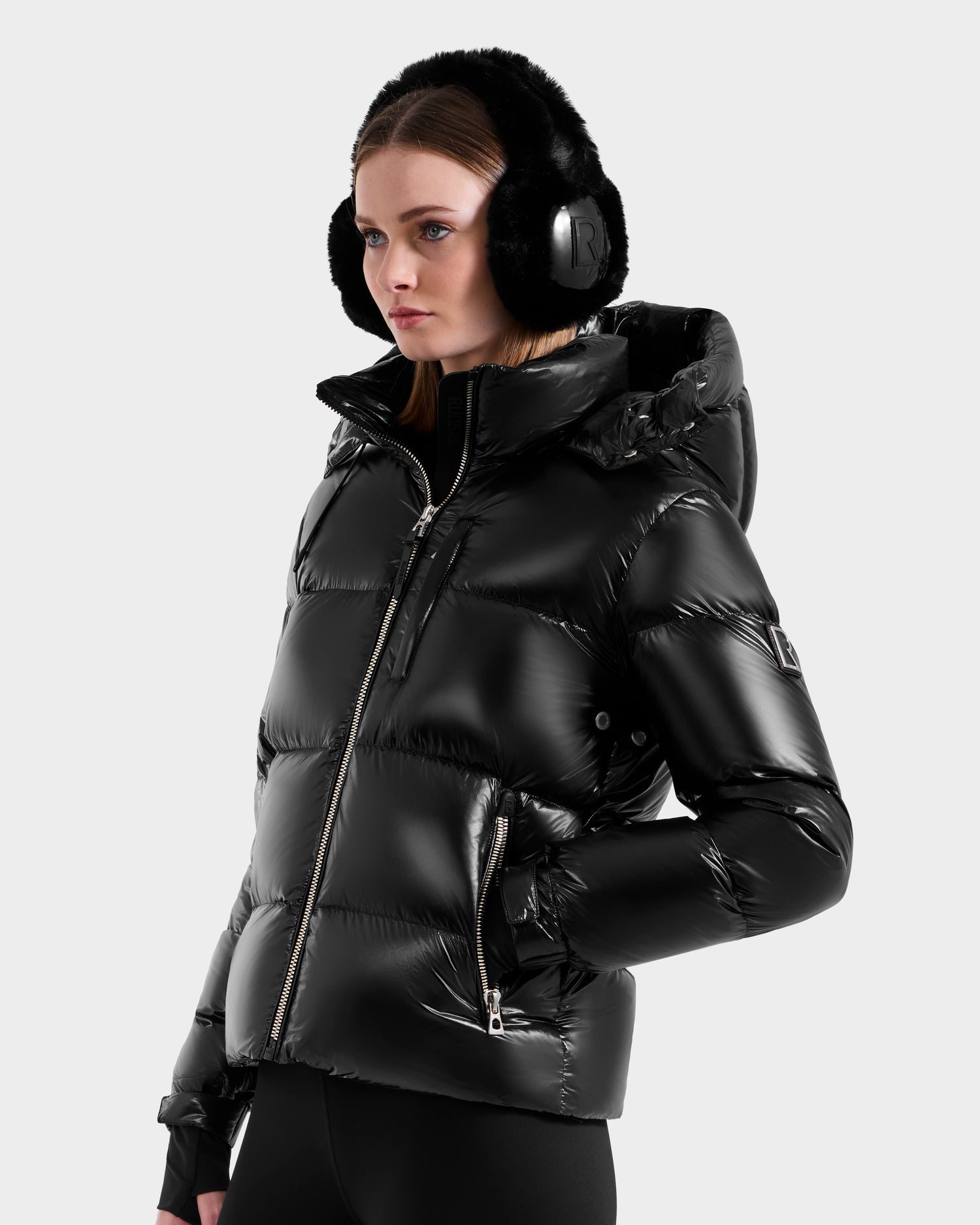 A woman wears the Rudsak ASHLYN S - 8124565 Black down puffer jacket with a hood, paired with black earmuffs and pants, standing against a light gray background and looking slightly to the side.