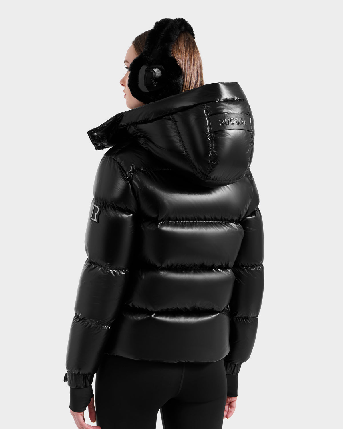 A person wearing black earmuffs and the Rudsak ASHLYN S - 8124565 Black shiny, water-resistant puffer jacket with a hood stands with their back to a plain light gray background.