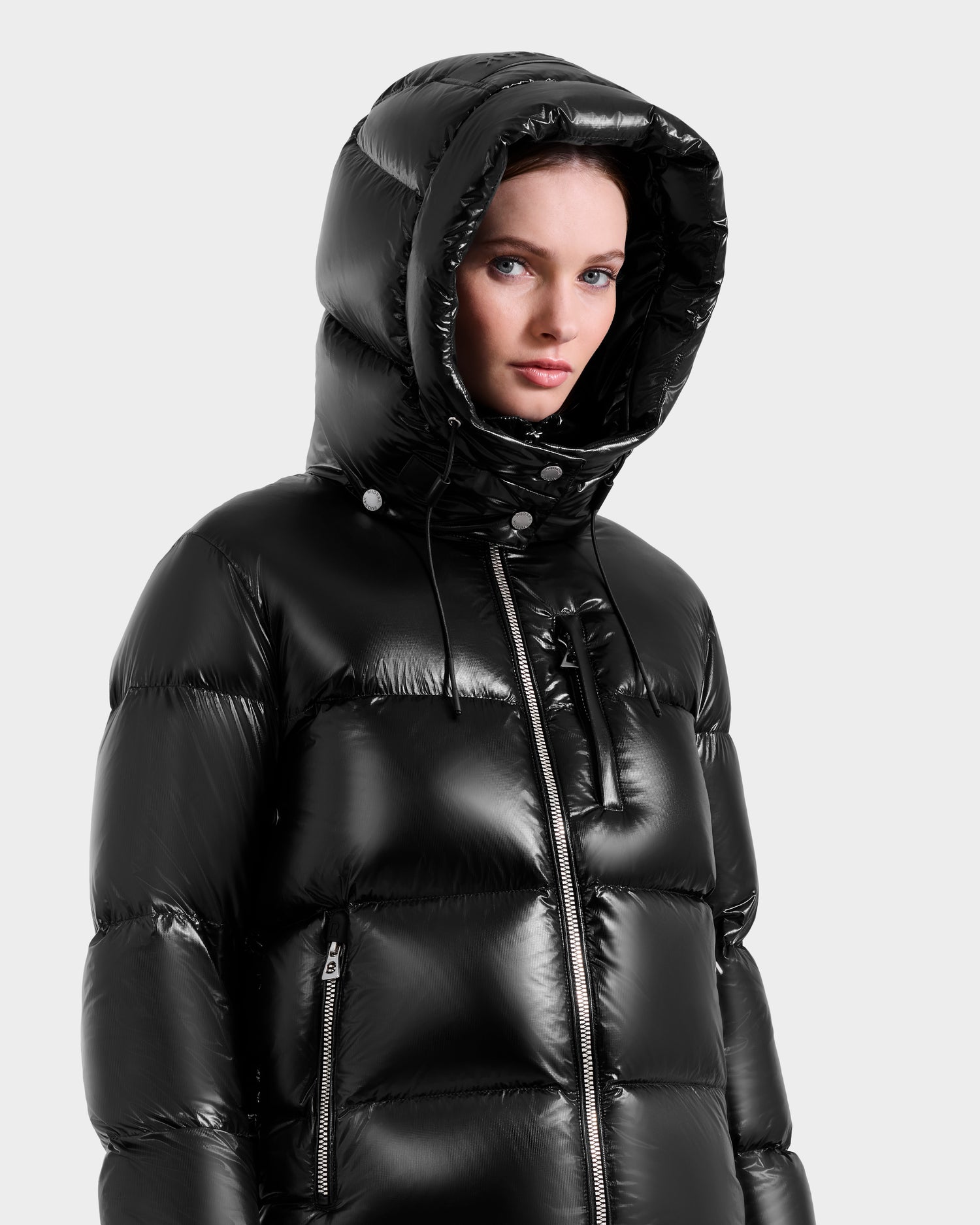A woman in the Rudsak ASHLYN S - 8124565 Black, an RDS-certified shiny black down puffer jacket with hood up, gazes slightly to the side against a plain light background.