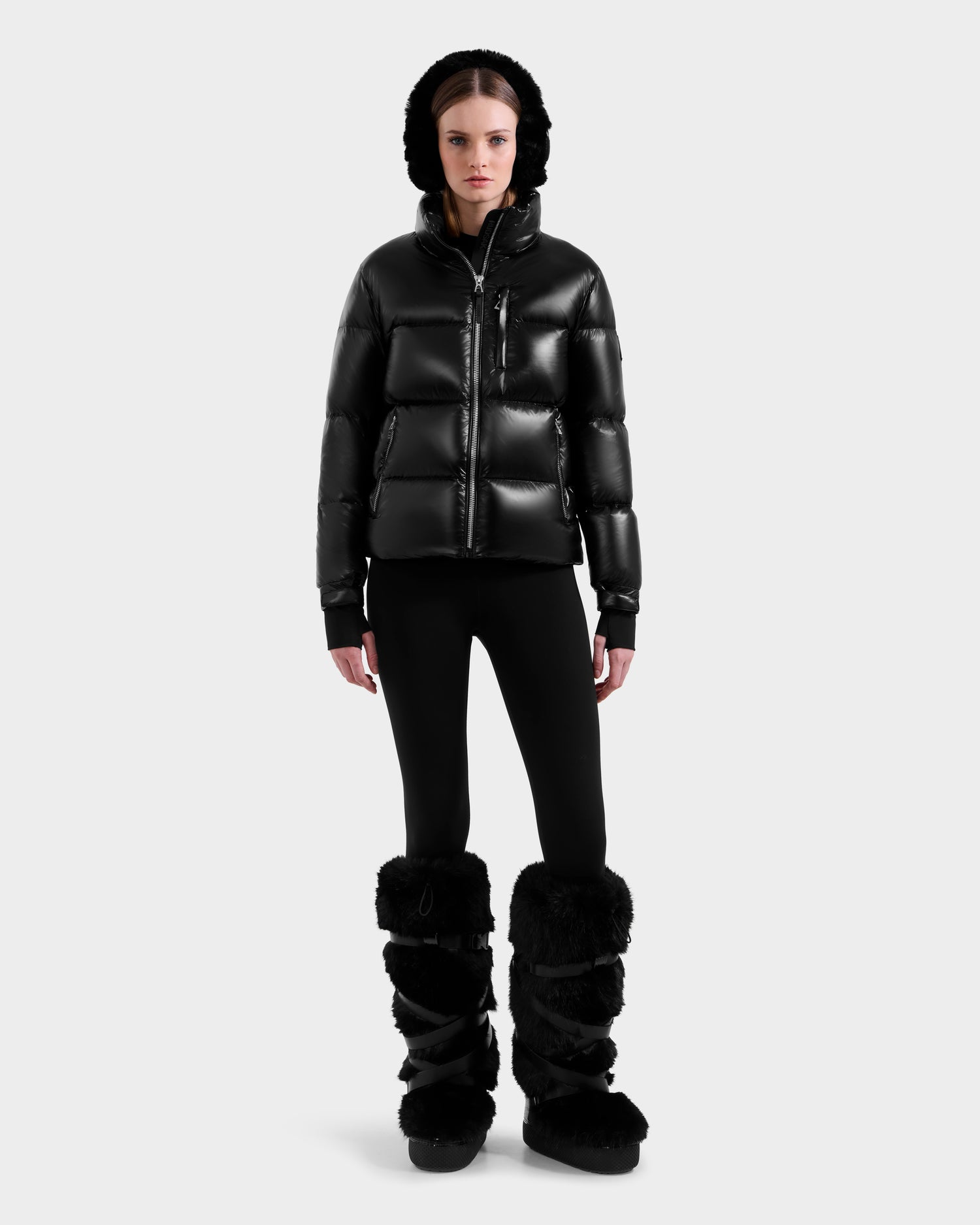 A person wearing the Rudsak ASHLYN S - 8124565 Black water-resistant puffer jacket with a hood, black leggings, and tall black furry boots stands facing forward against a plain light background.