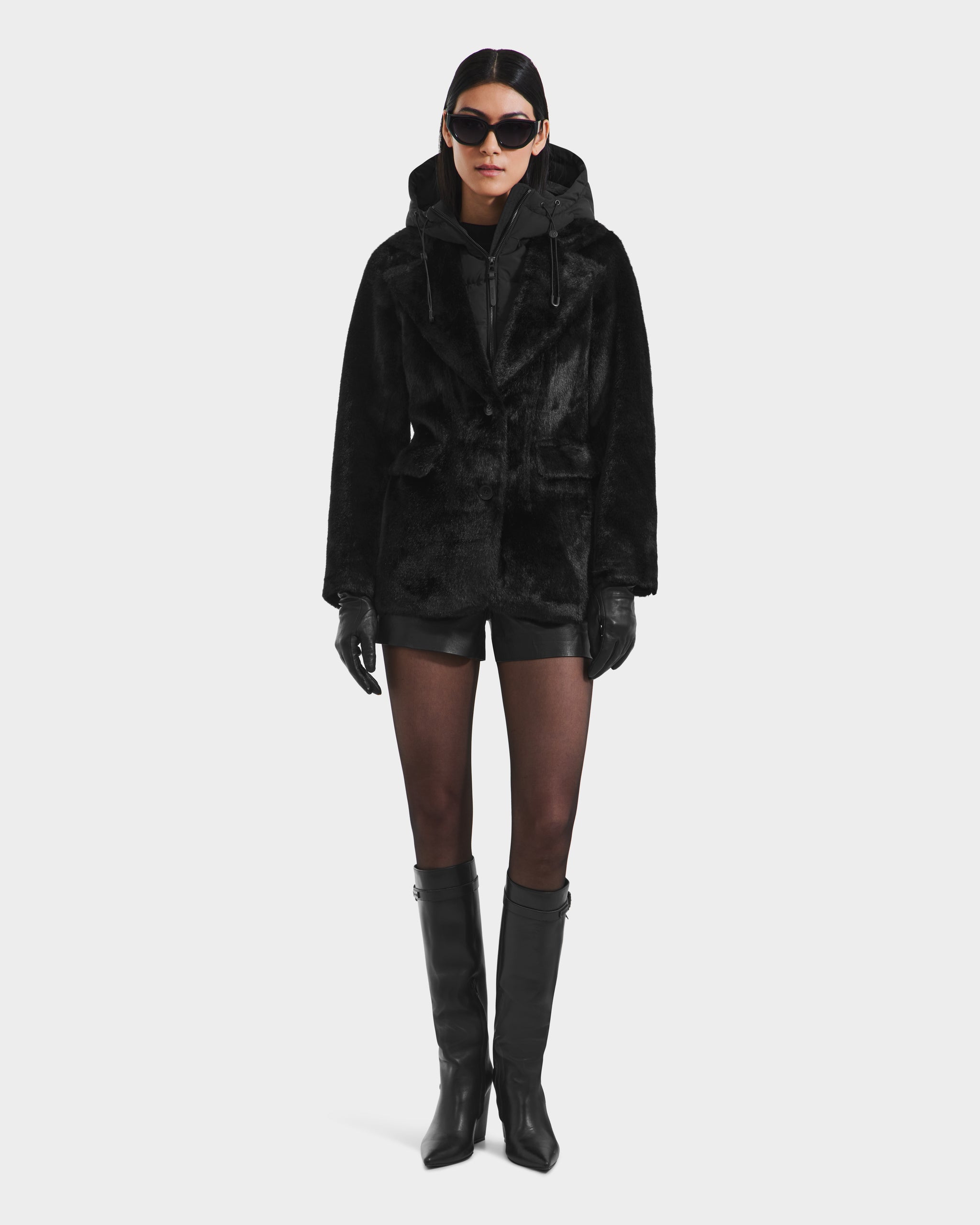 A person wearing the Rudsak ASPEN F - 8125604 Black women's faux fur jacket with removable hooded dickie, black shorts, sheer tights, knee-high boots, gloves, and large sunglasses stands facing forward against a plain light background.