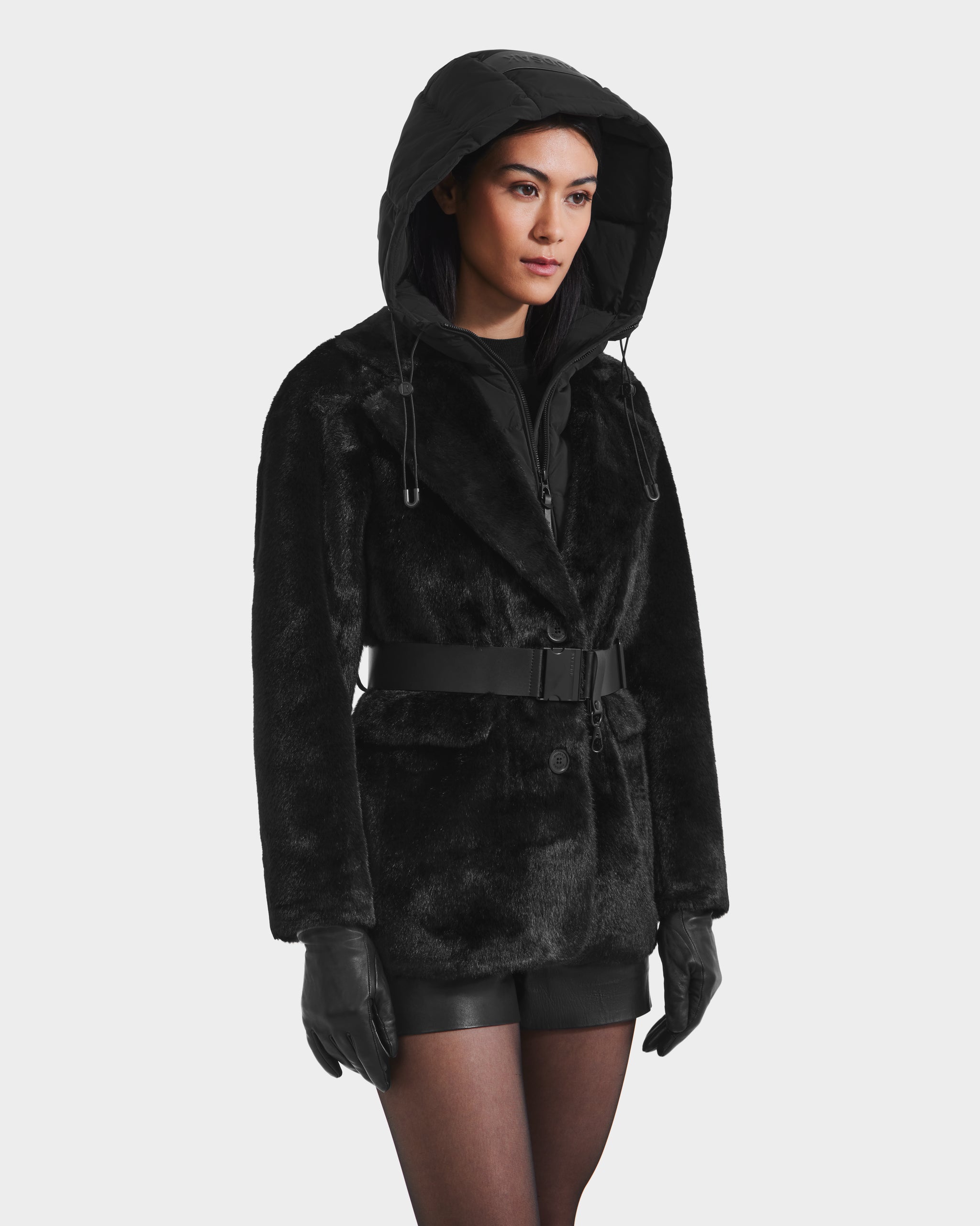 A woman wears the Rudsak ASPEN F - 8125604 Black faux fur jacket with belted waist and removable hooded dickie, paired with black gloves, shorts, and sheer tights—an effortlessly chic choice for cold-weather style.