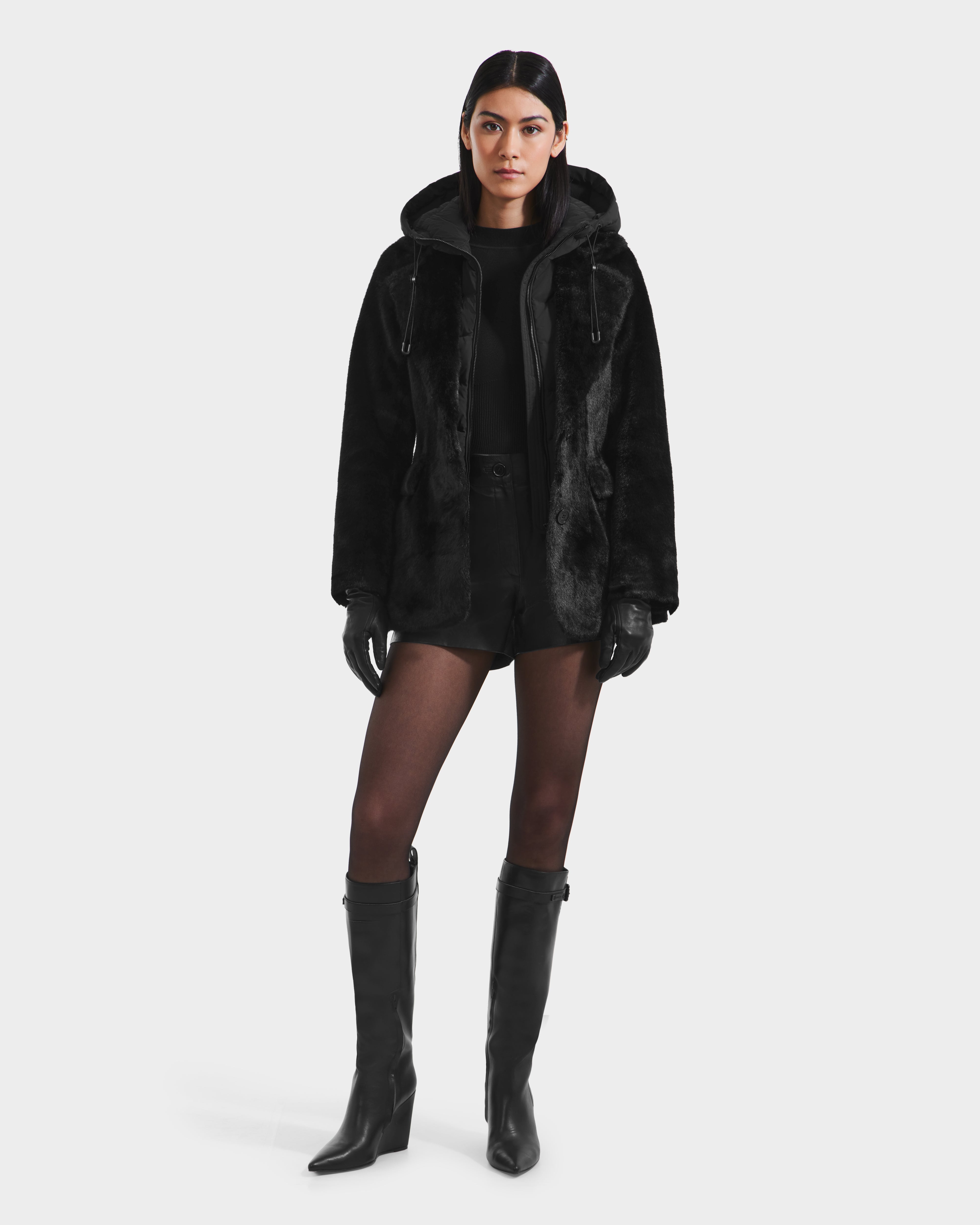 A woman faces forward in the Rudsak ASPEN F - 8125604 Black faux fur jacket with a removable hooded dickie, styled with black gloves, shorts, sheer tights, and knee-high boots against a plain white background.