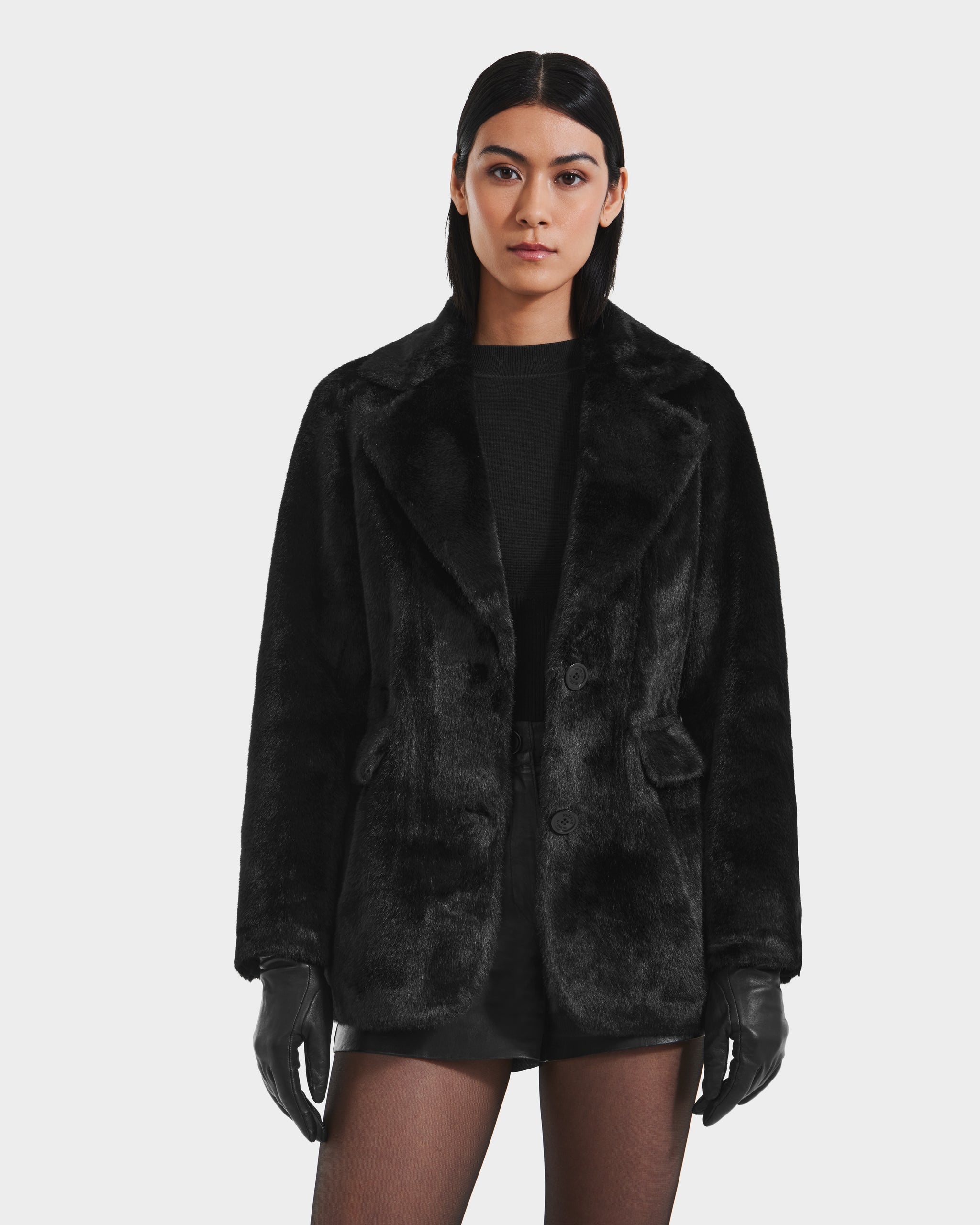 A woman with straight, dark hair wears the Rudsak ASPEN F - 8125604 Black women's faux fur jacket with a removable hooded dickie, paired with black gloves, a black top, shorts, and sheer tights against a white background.