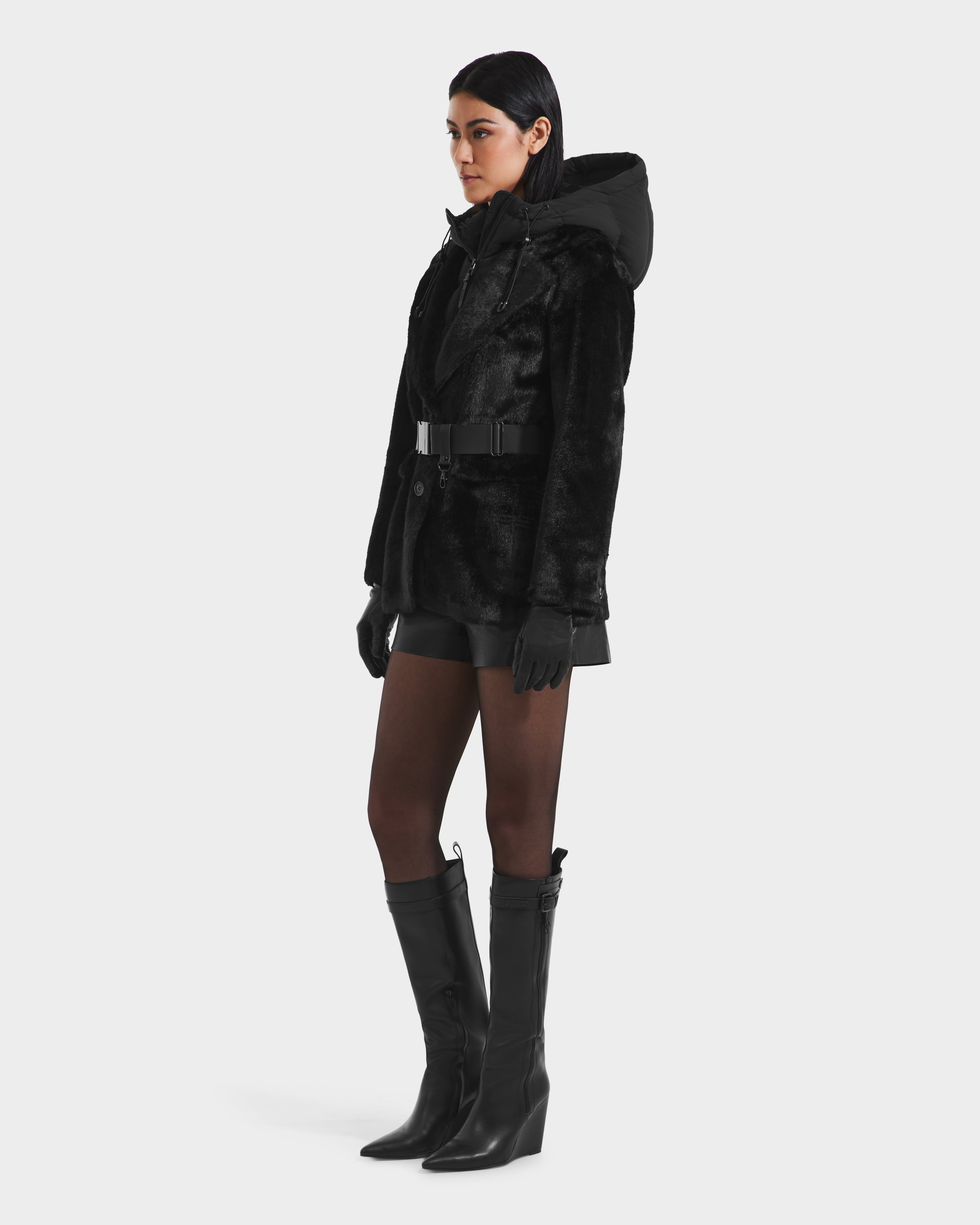 A woman faces left against a white background, wearing the Rudsak ASPEN F - 8125604 Black women’s faux fur jacket with removable hooded dickie—perfect for any cold-weather wardrobe.