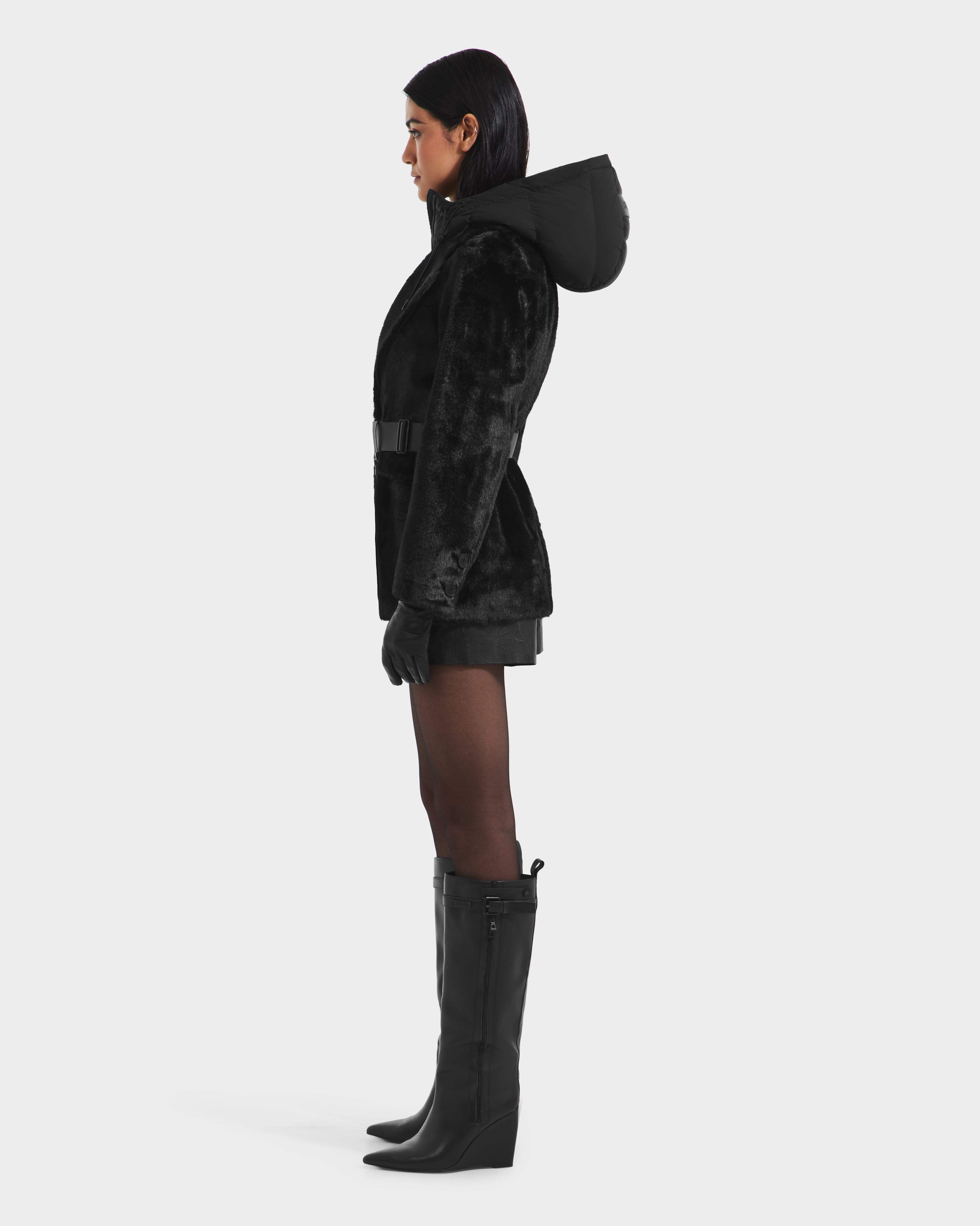 A woman stands in profile wearing the Rudsak ASPEN F - 8125604 Black jacket, styled with gloves, a short skirt or shorts, sheer black tights, and knee-high boots against a plain light background.