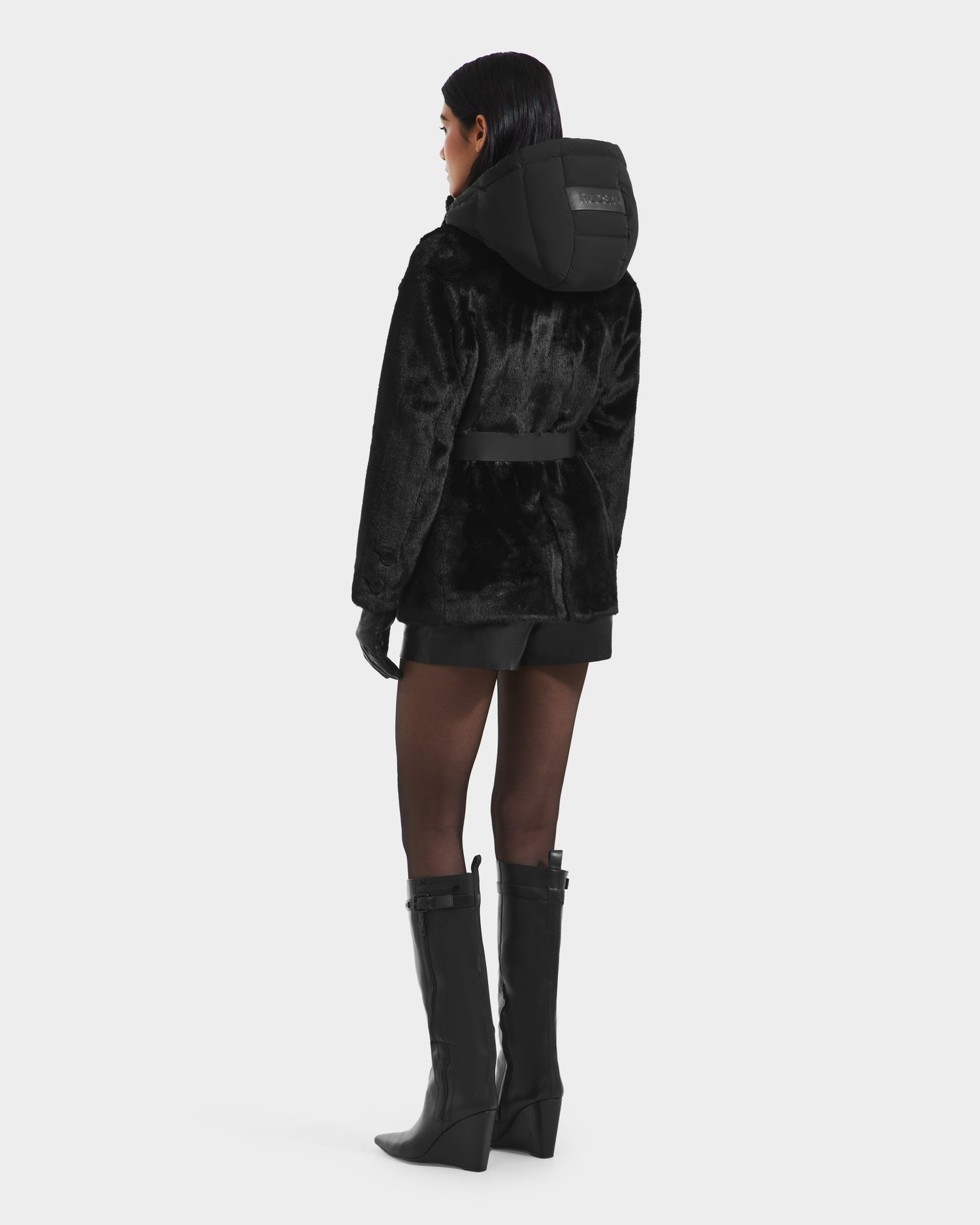 A woman faces away wearing the Rudsak ASPEN F - 8125604 Black faux fur jacket with a belted waist and hood, paired with black gloves, shorts, and knee-high boots—a chic, cold-weather look. The jacket sports a branded label on the hood.