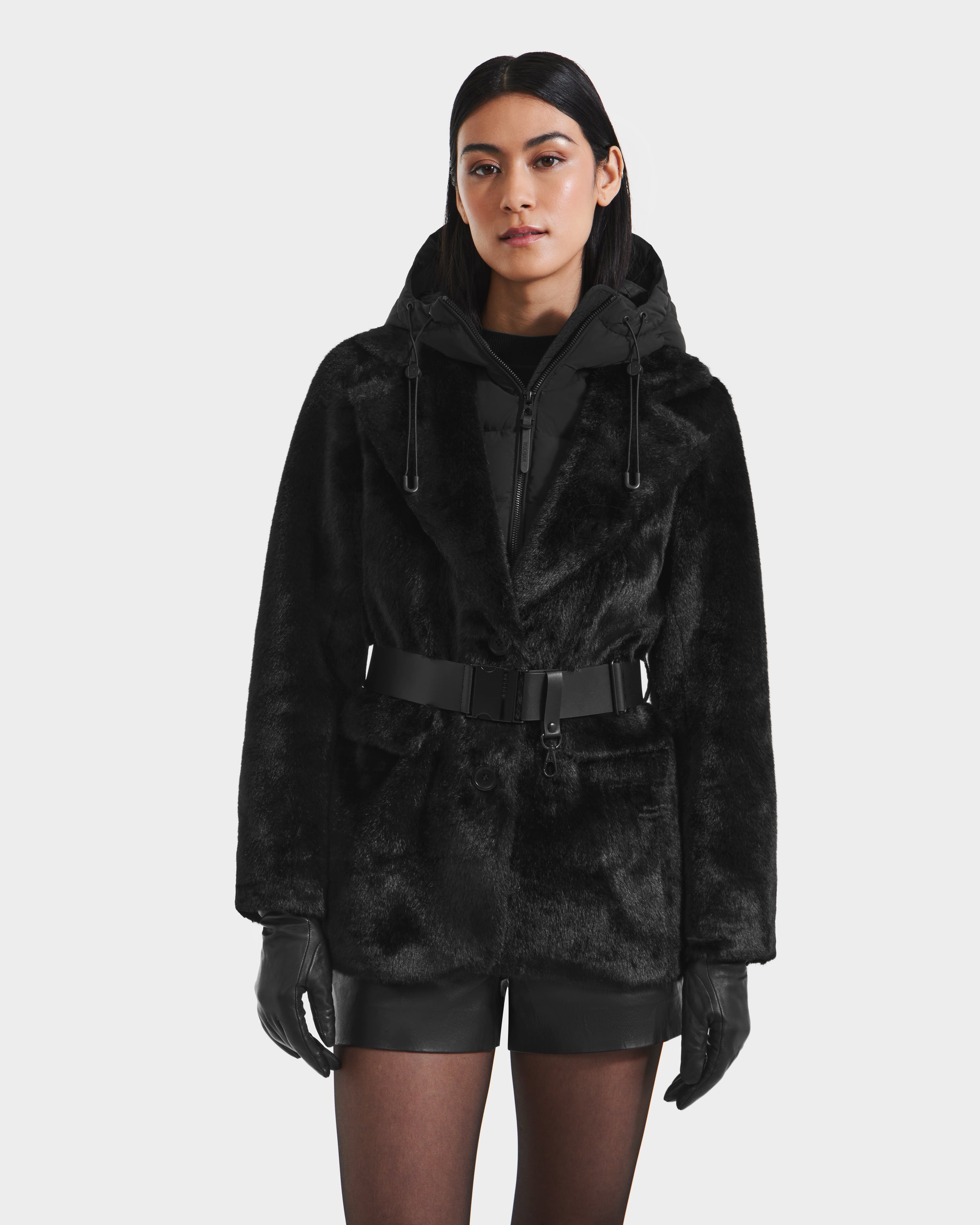 A woman models the Rudsak ASPEN F - 8125604 Black faux fur jacket with belted waist and removable hooded dickie, styled with black gloves, shorts, and sheer tights against a white background—ideal for chic winter looks.