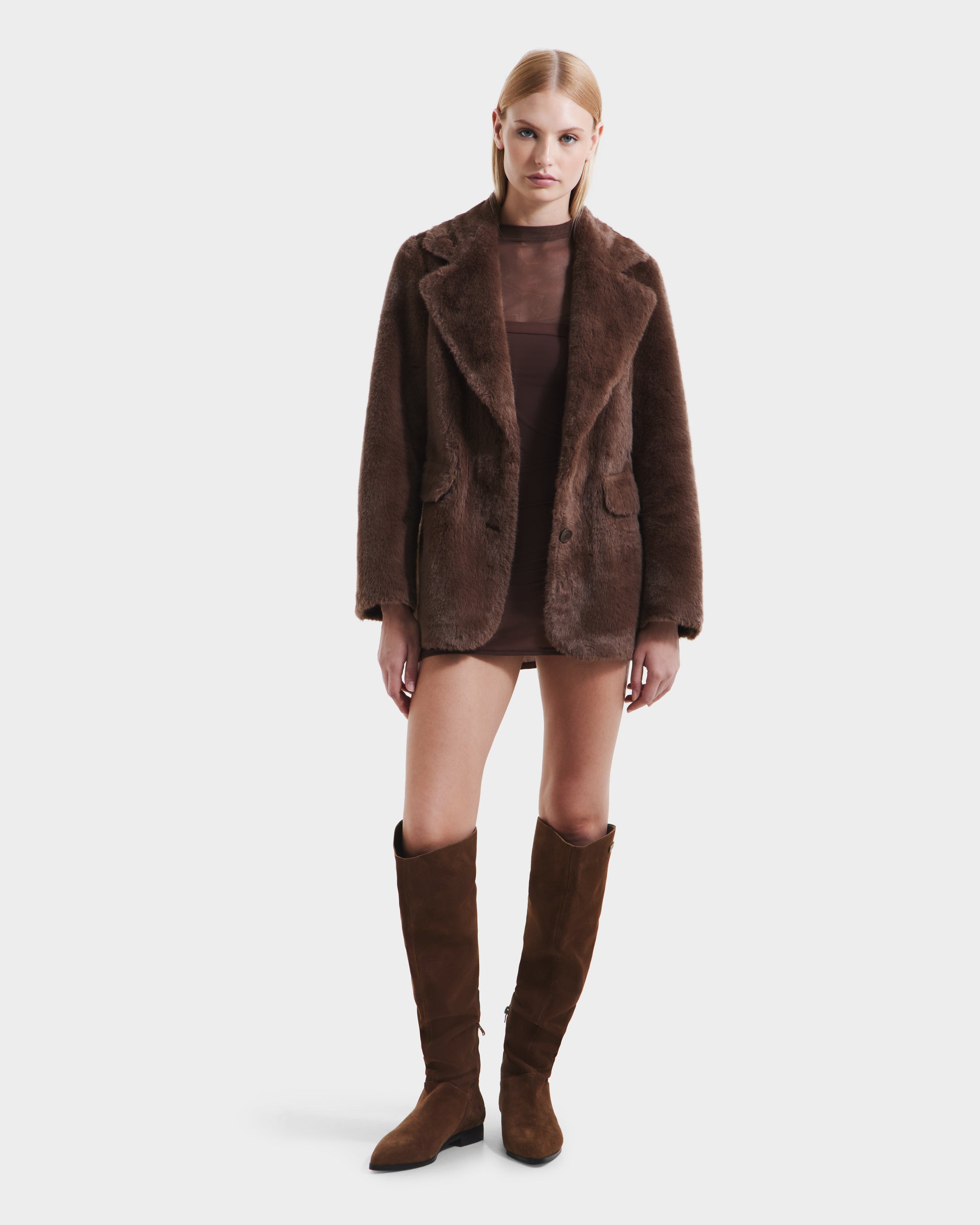 A woman faces forward in a Rudsak ASPEN F - 8125604 Mocha faux fur jacket, layered over a sheer brown dress with knee-high suede boots, showcasing a chic cold-weather look against a plain white background.