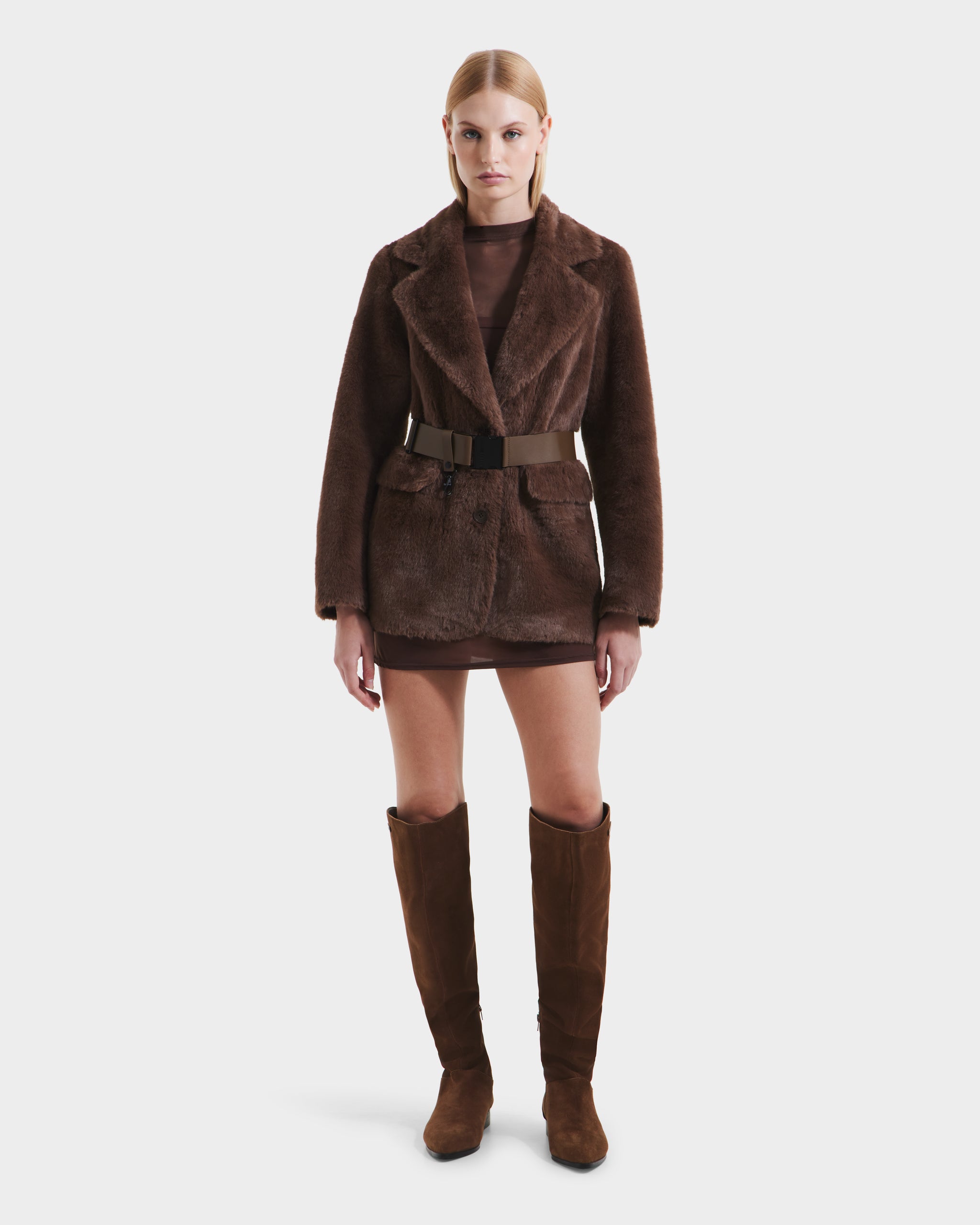 A woman faces forward in the Rudsak ASPEN F - 8125604 Mocha faux fur jacket with a wide belt, paired with a short brown dress and knee-high suede boots—an elevated cold-weather look—set against a plain white background.