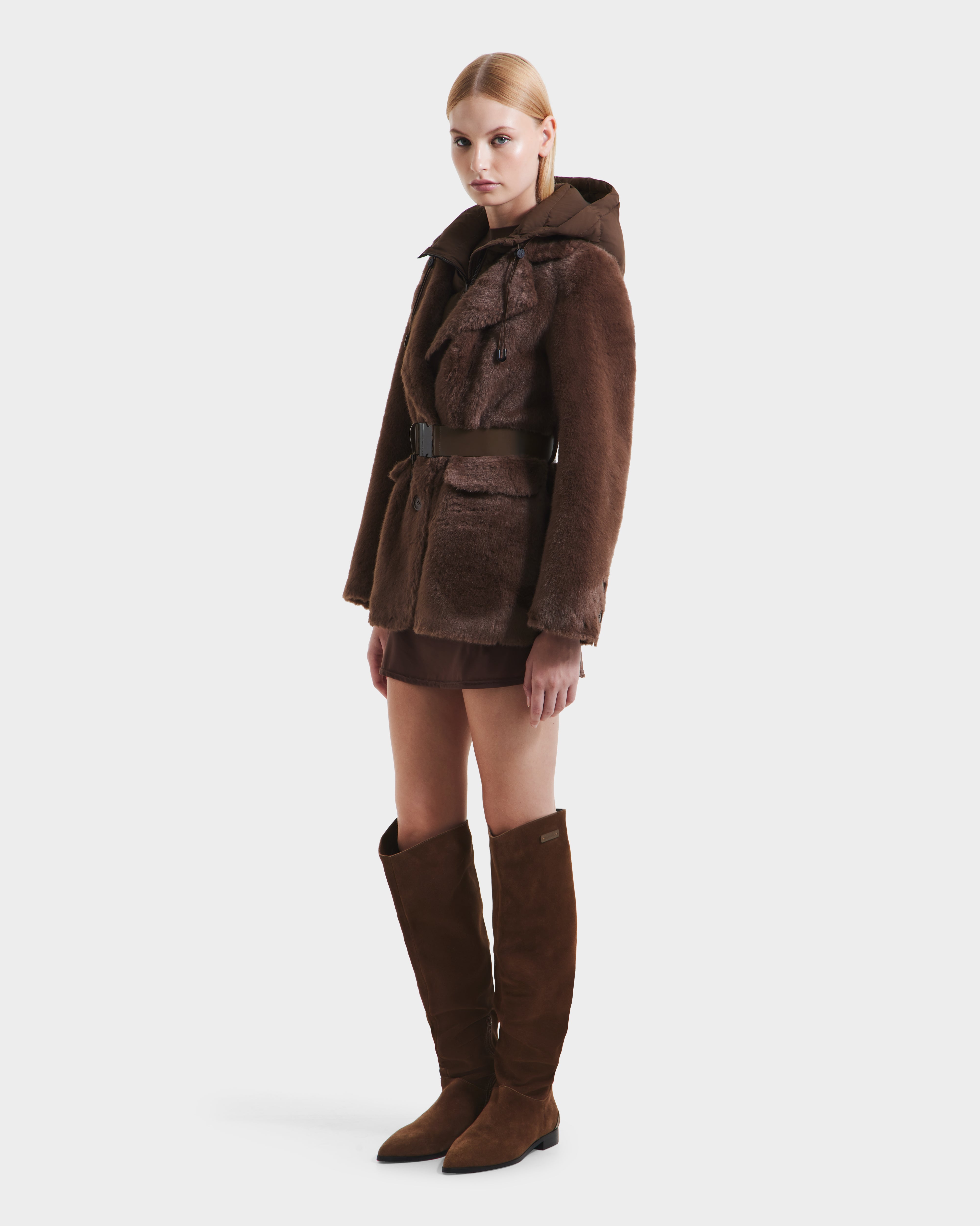 A blonde woman wears the Rudsak ASPEN F - 8125604 Mocha faux fur jacket with a belt, paired with a brown skirt and knee-high brown suede boots, perfect for cold weather, standing against a plain white background.