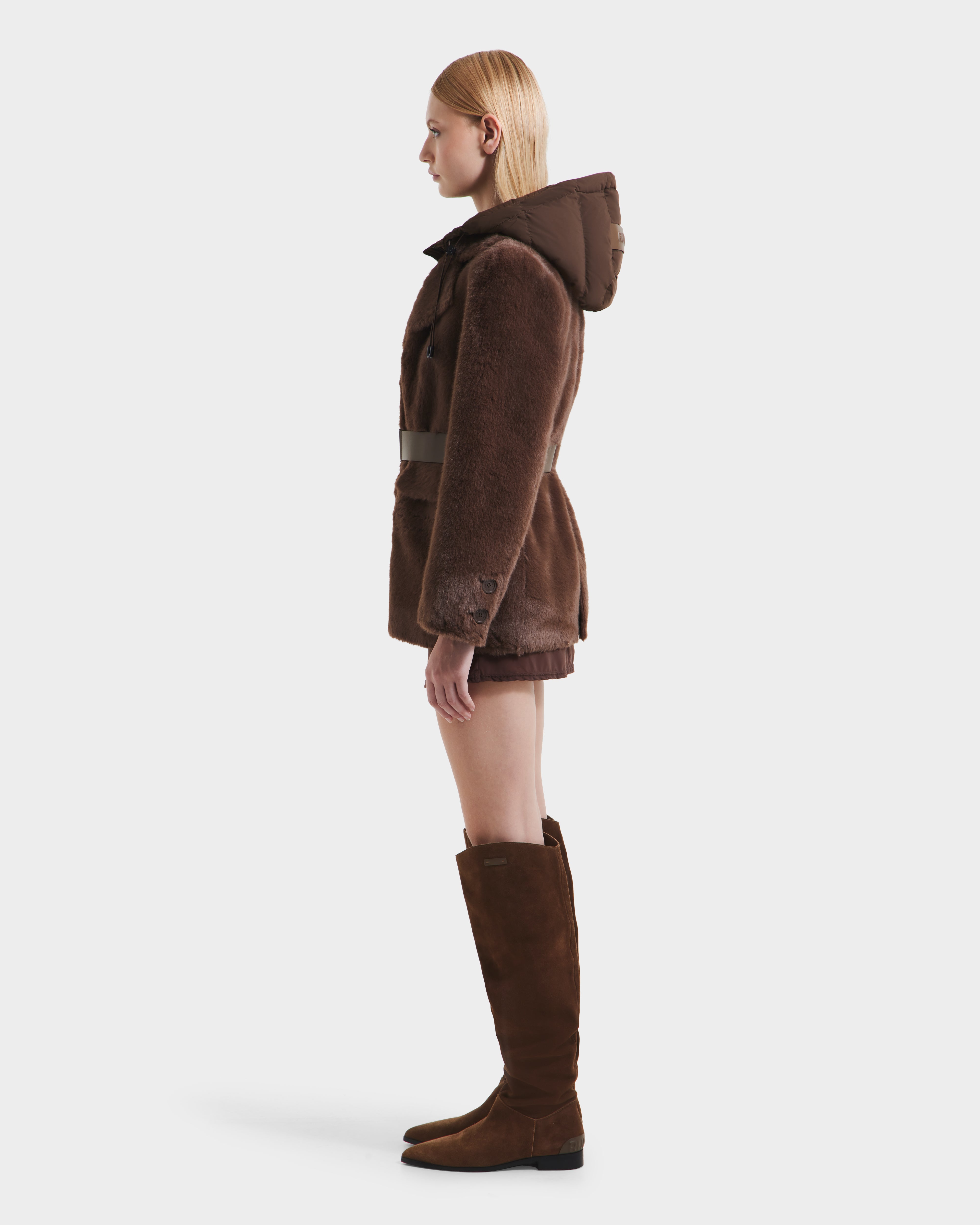 A woman with straight blonde hair faces left, modeling the Rudsak ASPEN F - 8125604 Mocha faux fur jacket with a removable down-insulated hood and tall brown suede boots, standing against a plain white background.