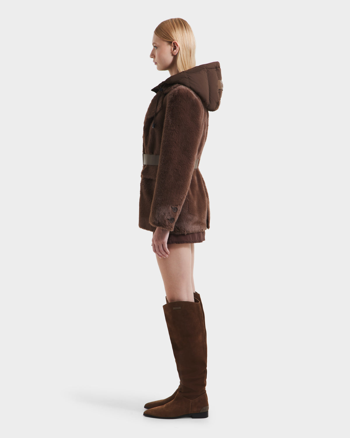 A woman with straight blonde hair faces left, modeling the Rudsak ASPEN F - 8125604 Mocha faux fur jacket with a removable down-insulated hood and tall brown suede boots, standing against a plain white background.