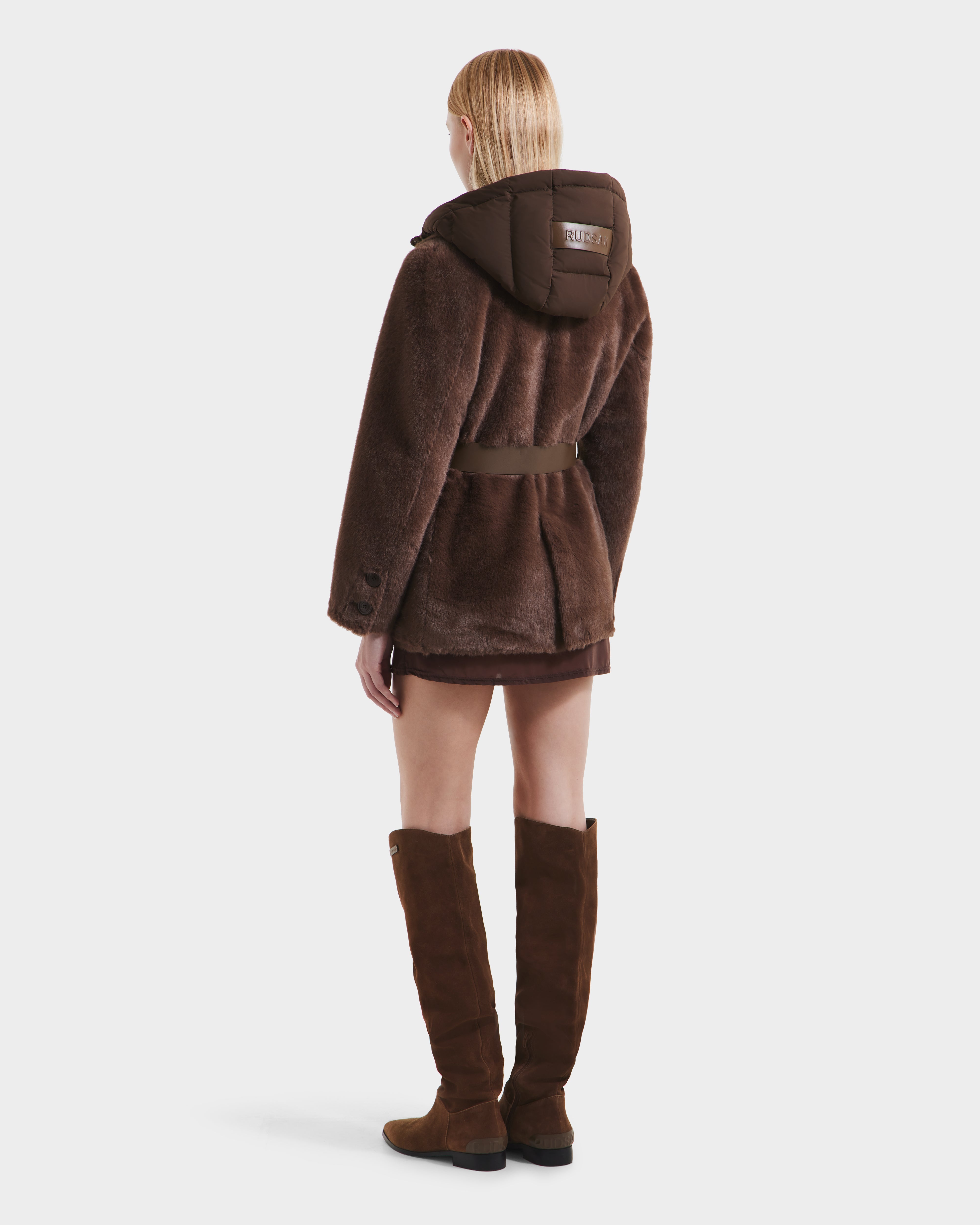 A person with straight blonde hair, seen from behind, models the Rudsak ASPEN F - 8125604 Mocha women's faux fur jacket with belted waist and removable hood, styled with a short skirt and knee-high brown suede boots against a plain white background.