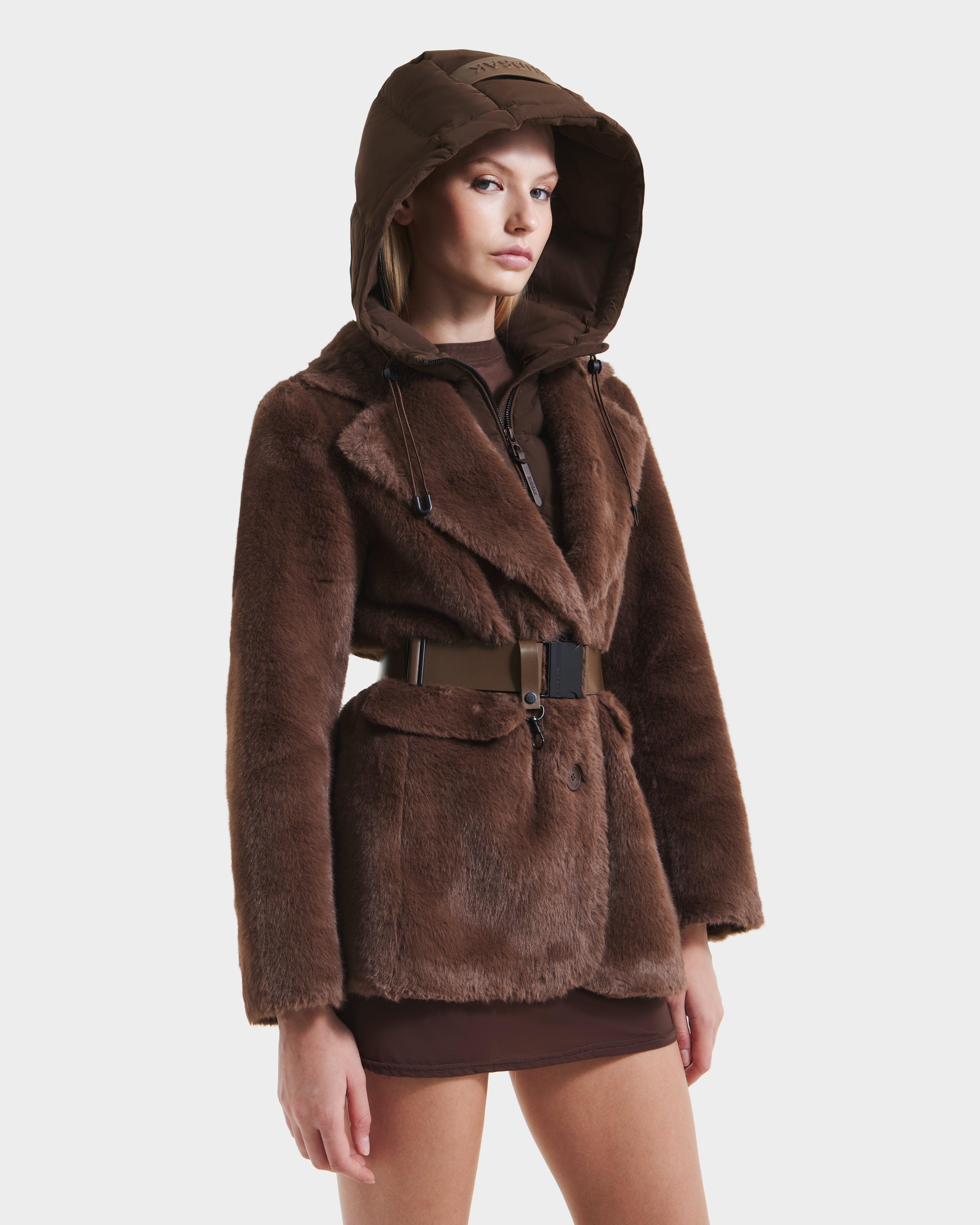 A woman models the Rudsak ASPEN F - 8125604 Mocha faux fur jacket with removable hooded dickie and wide belt over a brown mini skirt, posing against a plain white background.