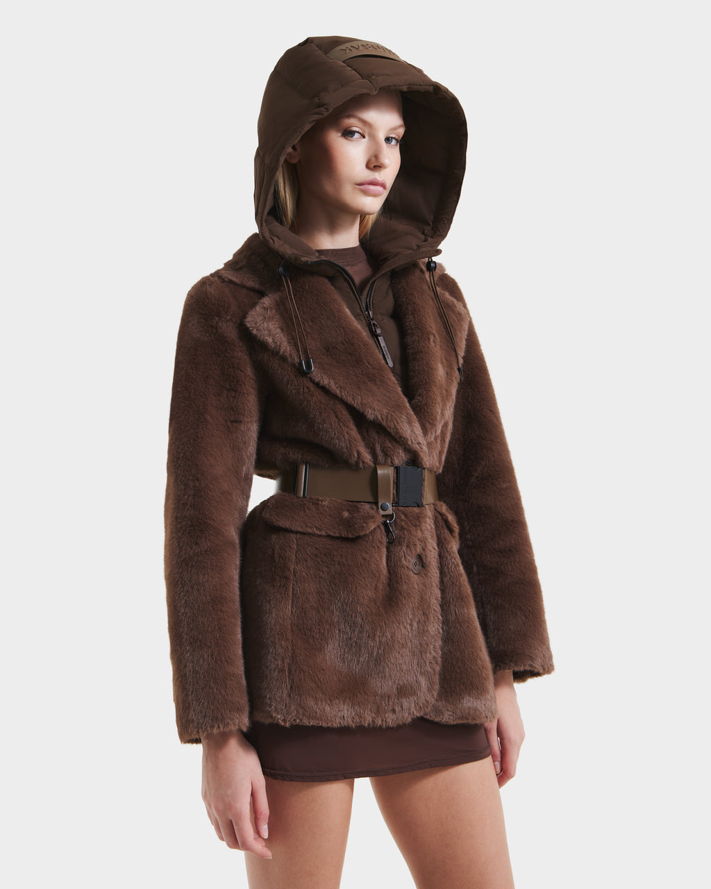 A woman models the Rudsak ASPEN F - 8125604 Mocha faux fur jacket with removable hooded dickie and wide belt over a brown mini skirt, posing against a plain white background.