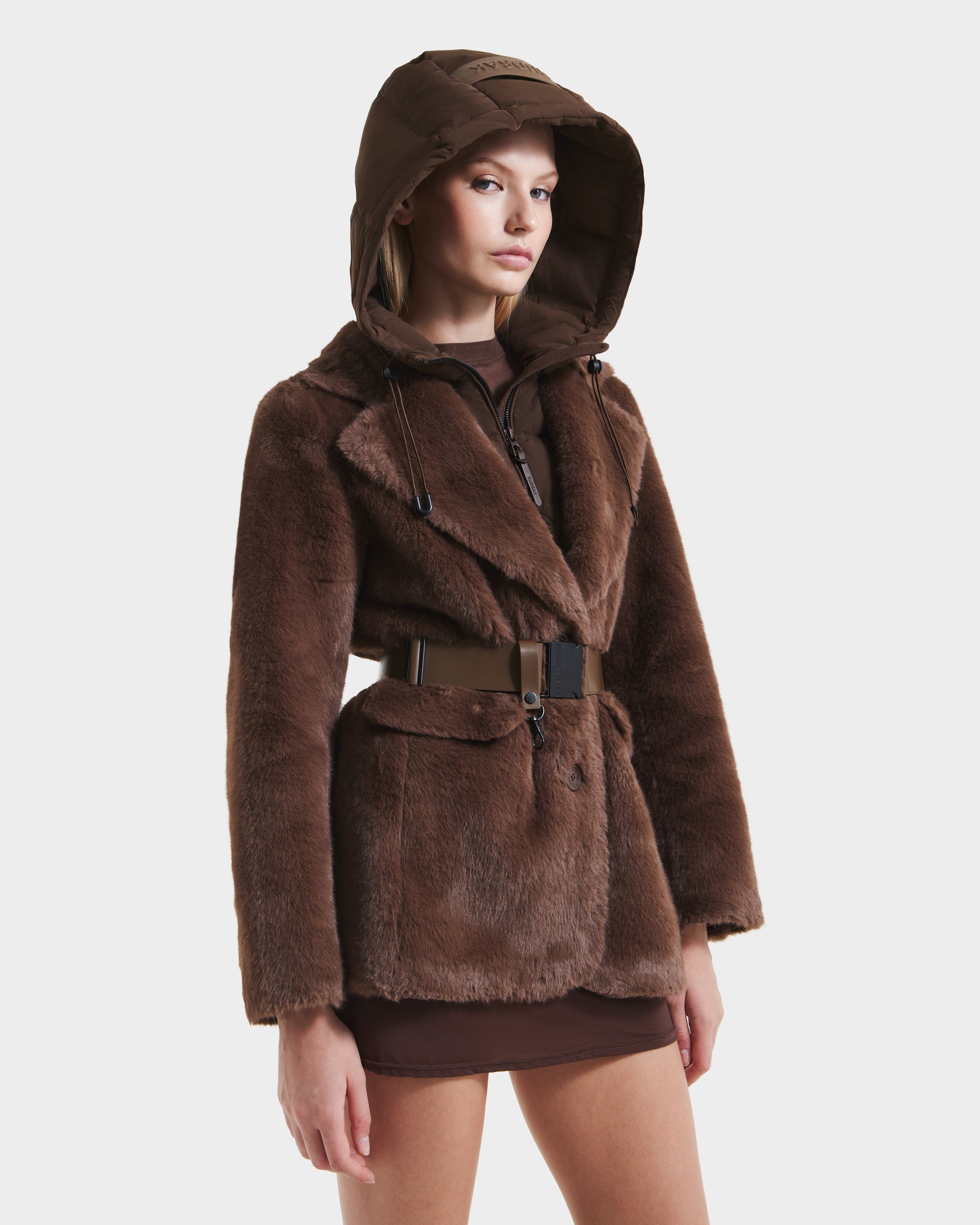A woman models the Rudsak ASPEN F - 8125604 Mocha faux fur jacket with removable hooded dickie and wide belt over a brown mini skirt, posing against a plain white background.