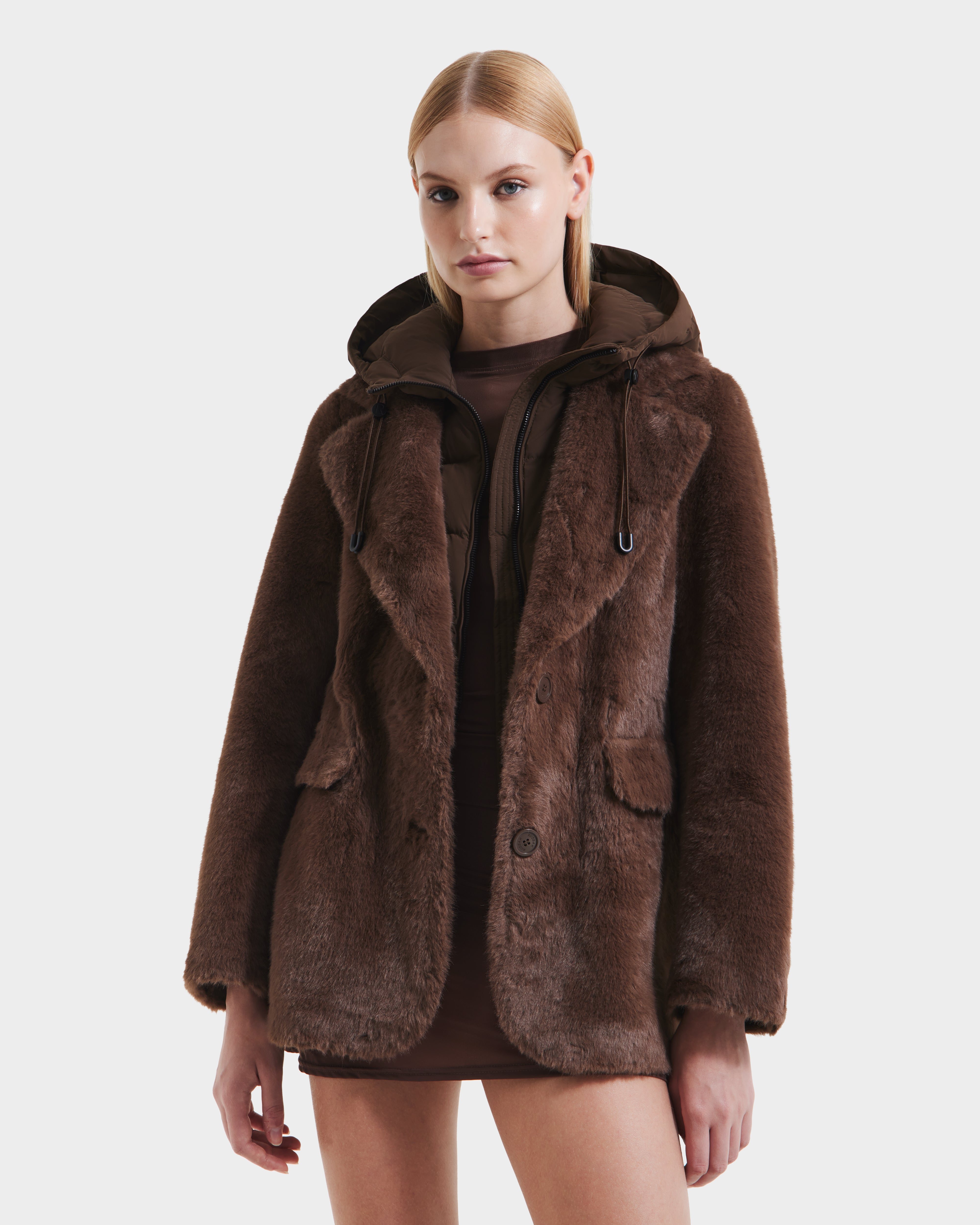 A woman with straight blonde hair models the Rudsak ASPEN F - 8125604 Mocha women’s faux fur jacket with a removable down-insulated hood over a short brown dress, standing against a plain white background.