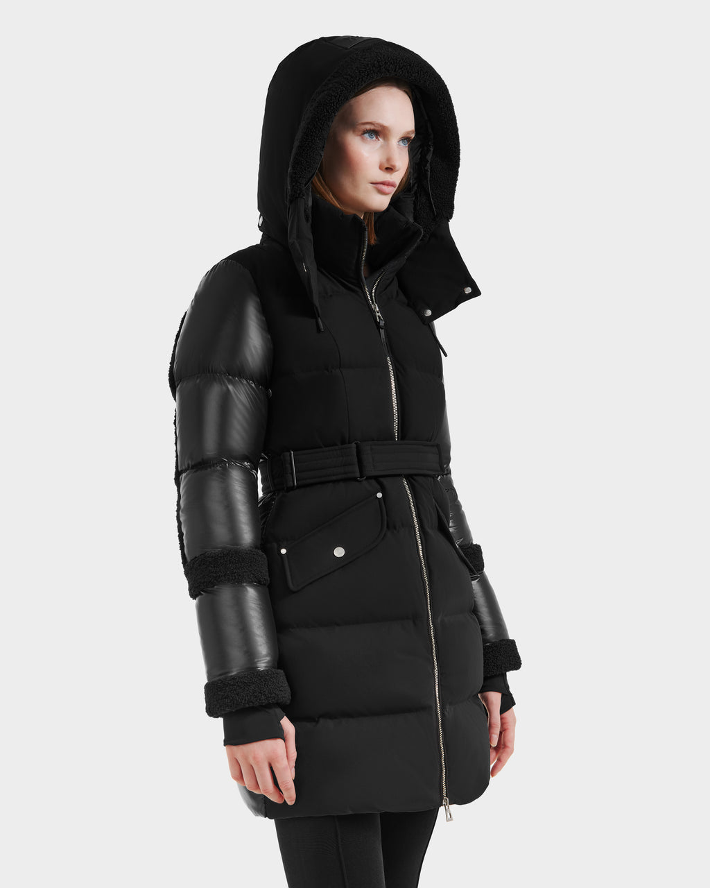 A woman wearing the Rudsak AUDREY L - 8124585 Black/Black knee-length down puffer with faux fur trim, shiny quilted sleeves, belted waist, and hood stands against a plain light gray background.