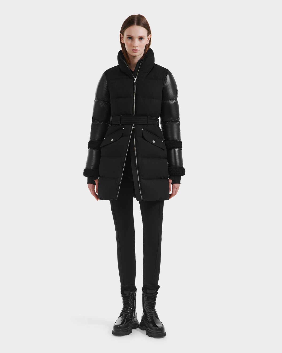 A person stands facing forward, wearing the Rudsak AUDREY L - 8124585 Black/Black knee-length down puffer jacket with zipper details, black pants, and black lace-up boots against a plain white background.